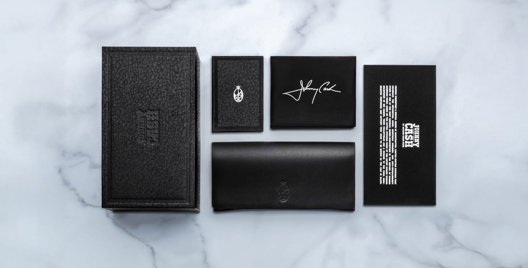 A flat lay of five black Jacques Marie Mage Cash items on white marble: a textured box, a small booklet, a card with white signature, a large case or wallet, and a black card with white text.