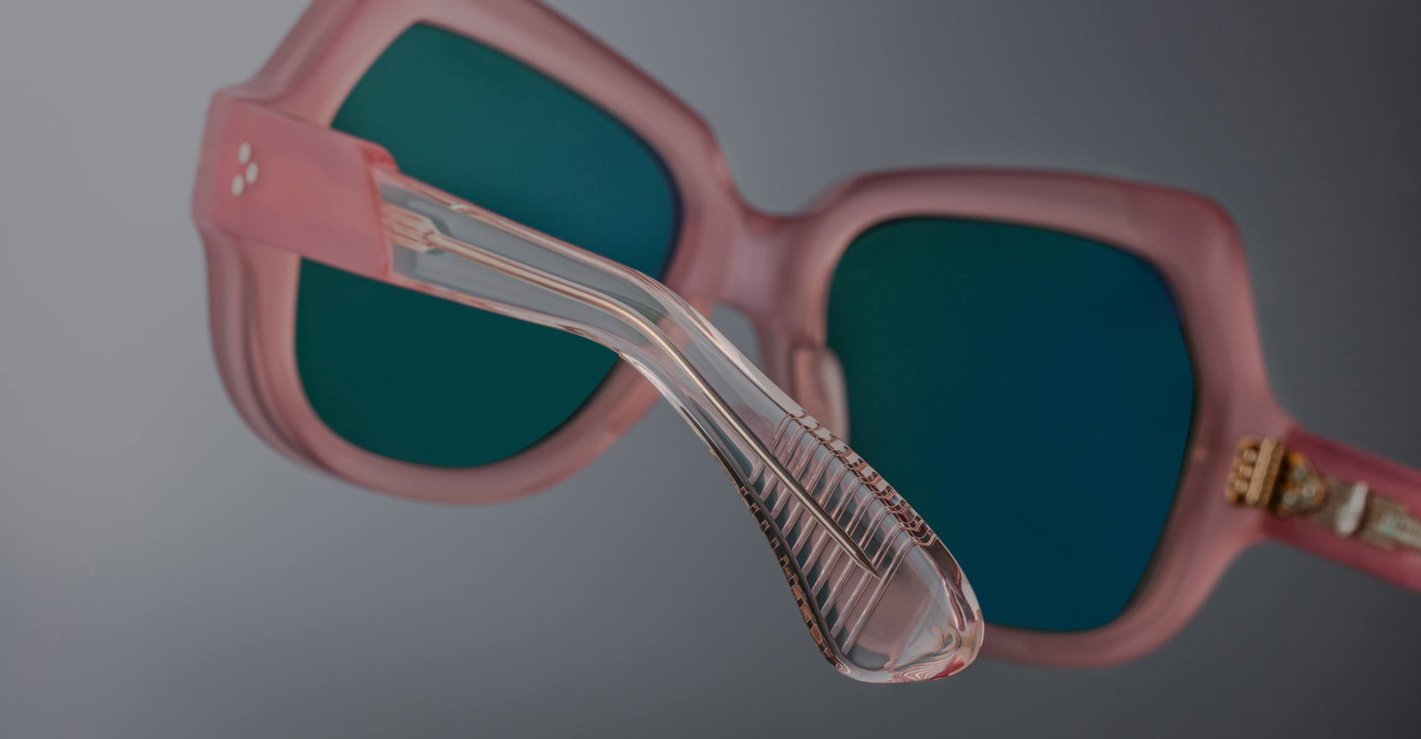Close-up of Jacques Marie Mage Perreti pink sunglasses with green tinted lenses, highlighting detailed texture on the transparent plastic arm against a gray background.