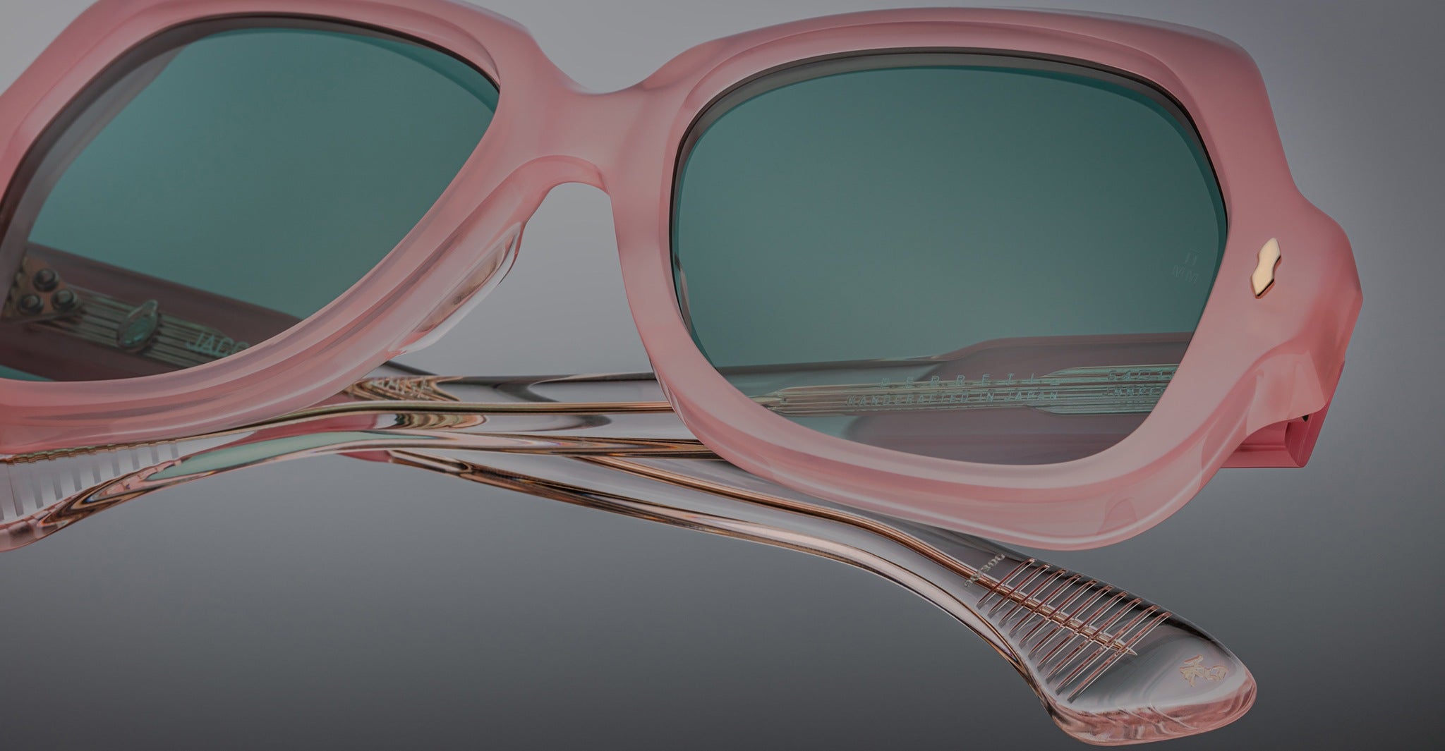 Close-up of Jacques Marie Mage Perreti sunglasses with pink frames and green-tinted lenses, highlighting detailed design on transparent, curved arms against a neutral gray background.