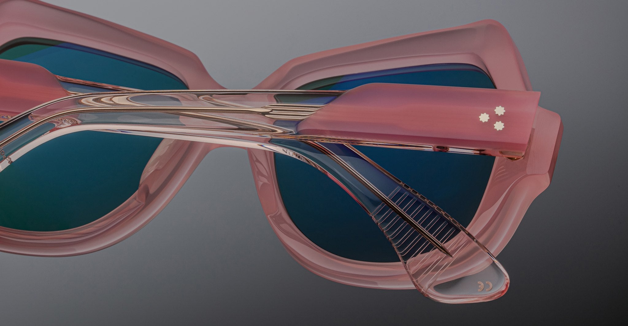 Close-up of Jacques Marie Mage Perreti sunglasses with stylish pink frames, blue-tinted lenses, and two small white star-shaped details on the temple, set against a neutral grey background.