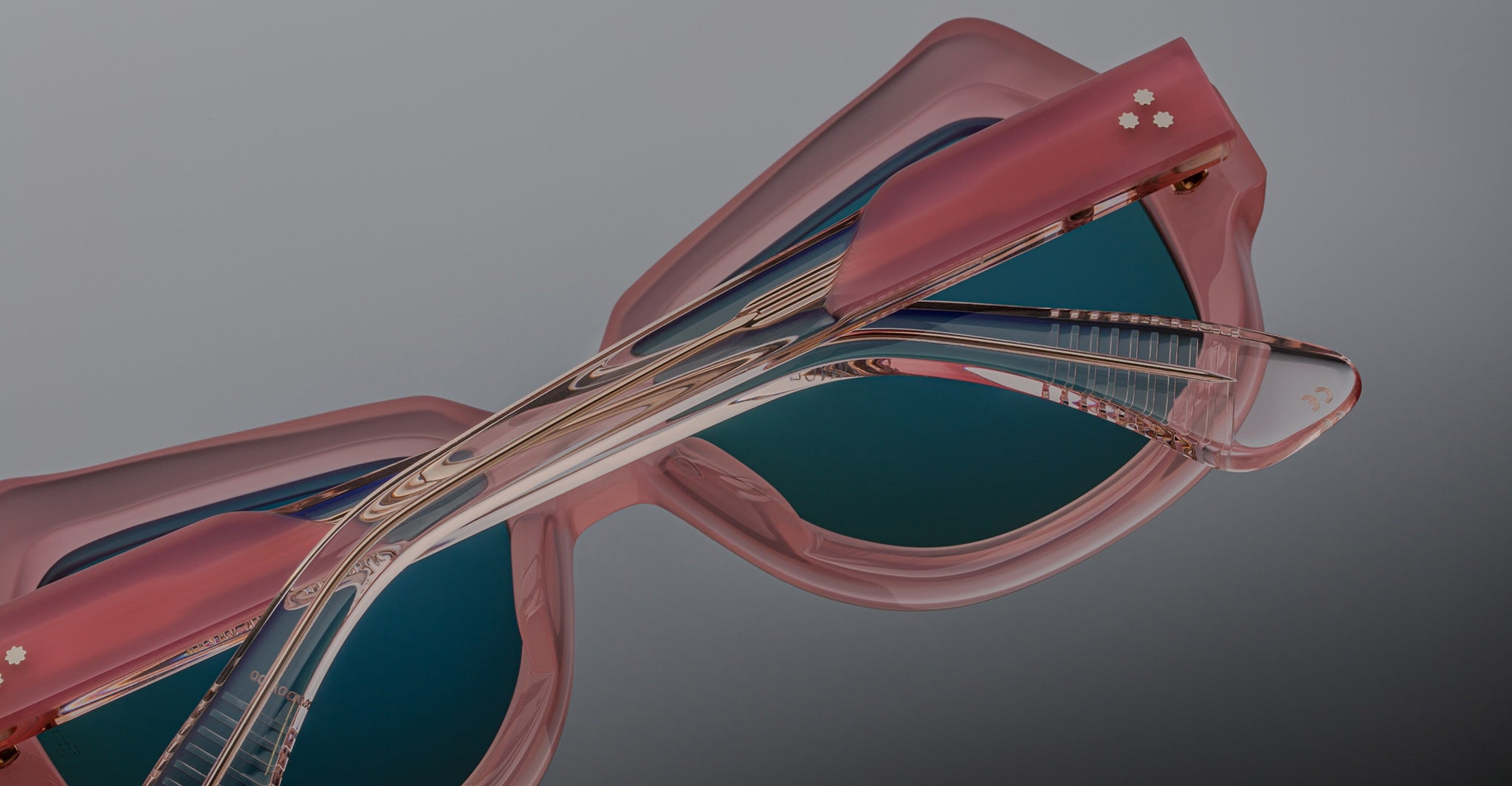 Close-up of Jacques Marie Mage Perreti pink sunglasses with thick frames and blue-tinted lenses, highlighting intricate details and reflections on the arms and lenses, set against a gray background.