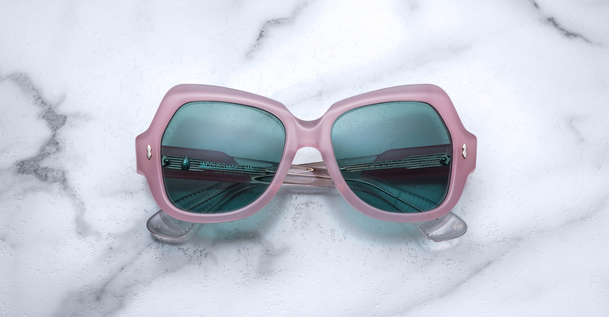 The Jacques Marie Mage Perreti sunglasses feature square pink frames and blue-tinted lenses, shown resting on a white marble surface.