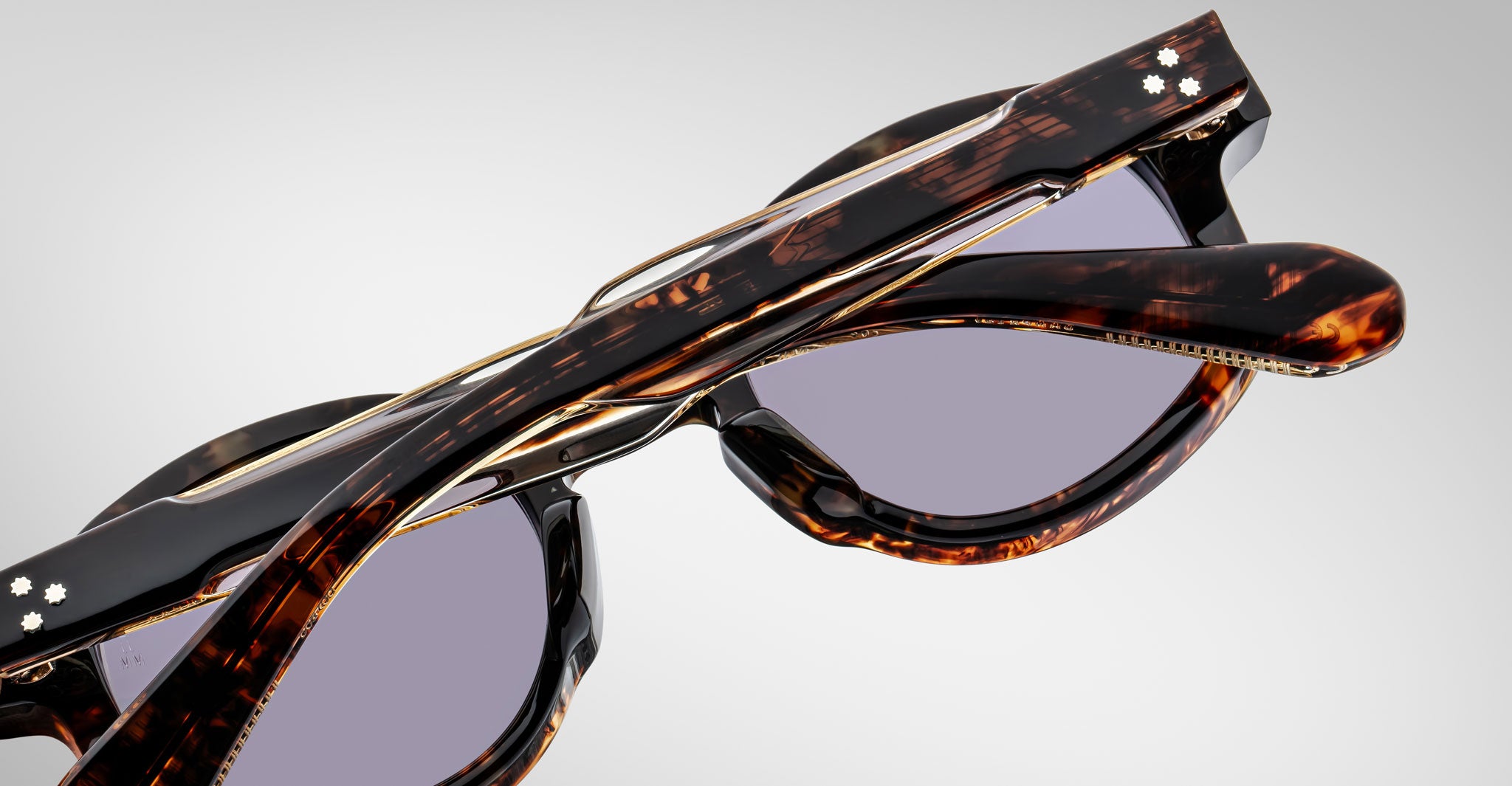 Close-up of two pairs of folded Jacques Marie Mage Petrus tortoiseshell sunglasses with purple-tinted lenses, stacked atop one another against a plain, light background.