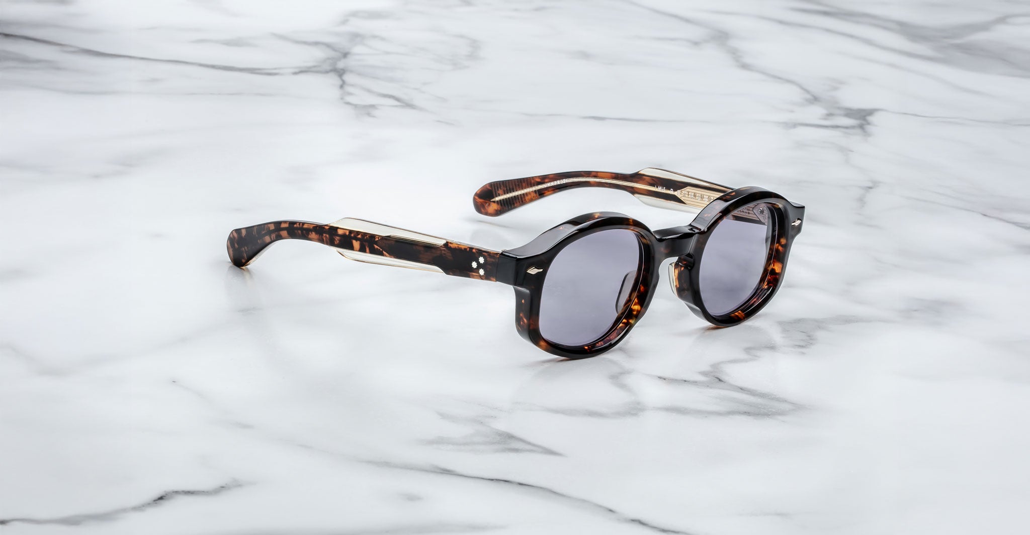 The Jacques Marie Mage Petrus round sunglasses with a tortoiseshell frame and dark lenses rest on a white marble surface, photographed from the side.