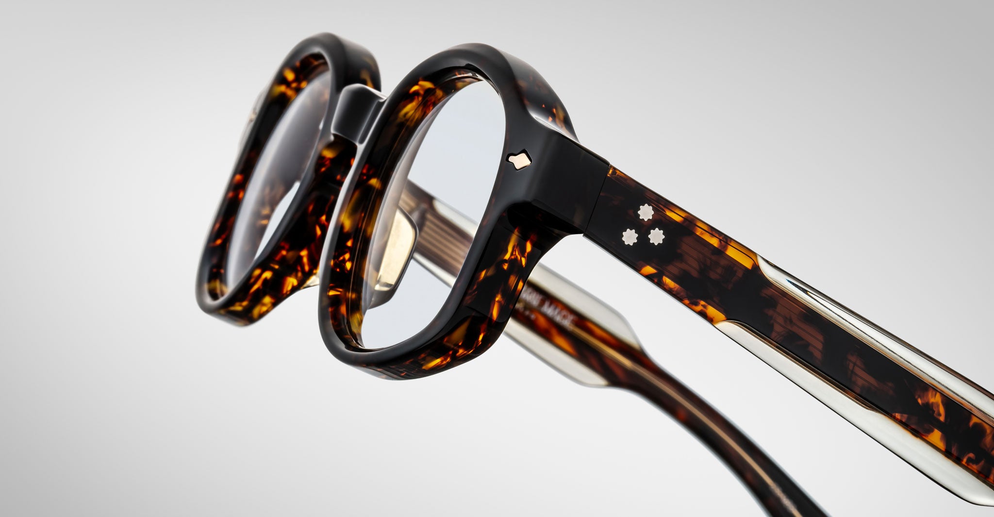 Close-up of Jacques Marie Mage Petrus eyeglasses with thick tortoiseshell frames, clear lenses, and partially visible arms featuring metal and decorative accents against a light gray background.