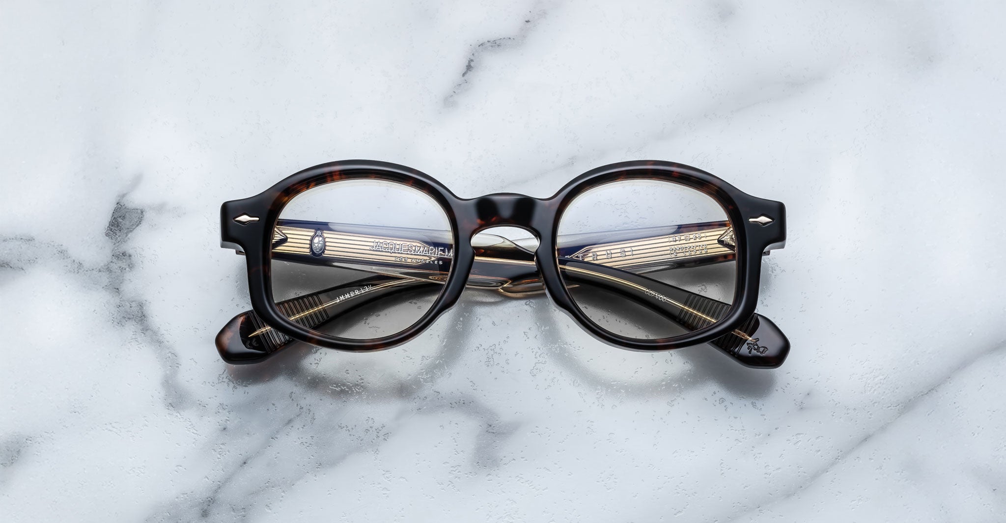 The Jacques Marie Mage Petrus eyeglasses, with round dark tortoise-shell frames and patterned arms, rest folded on a white marble surface, their details visible through the lenses.