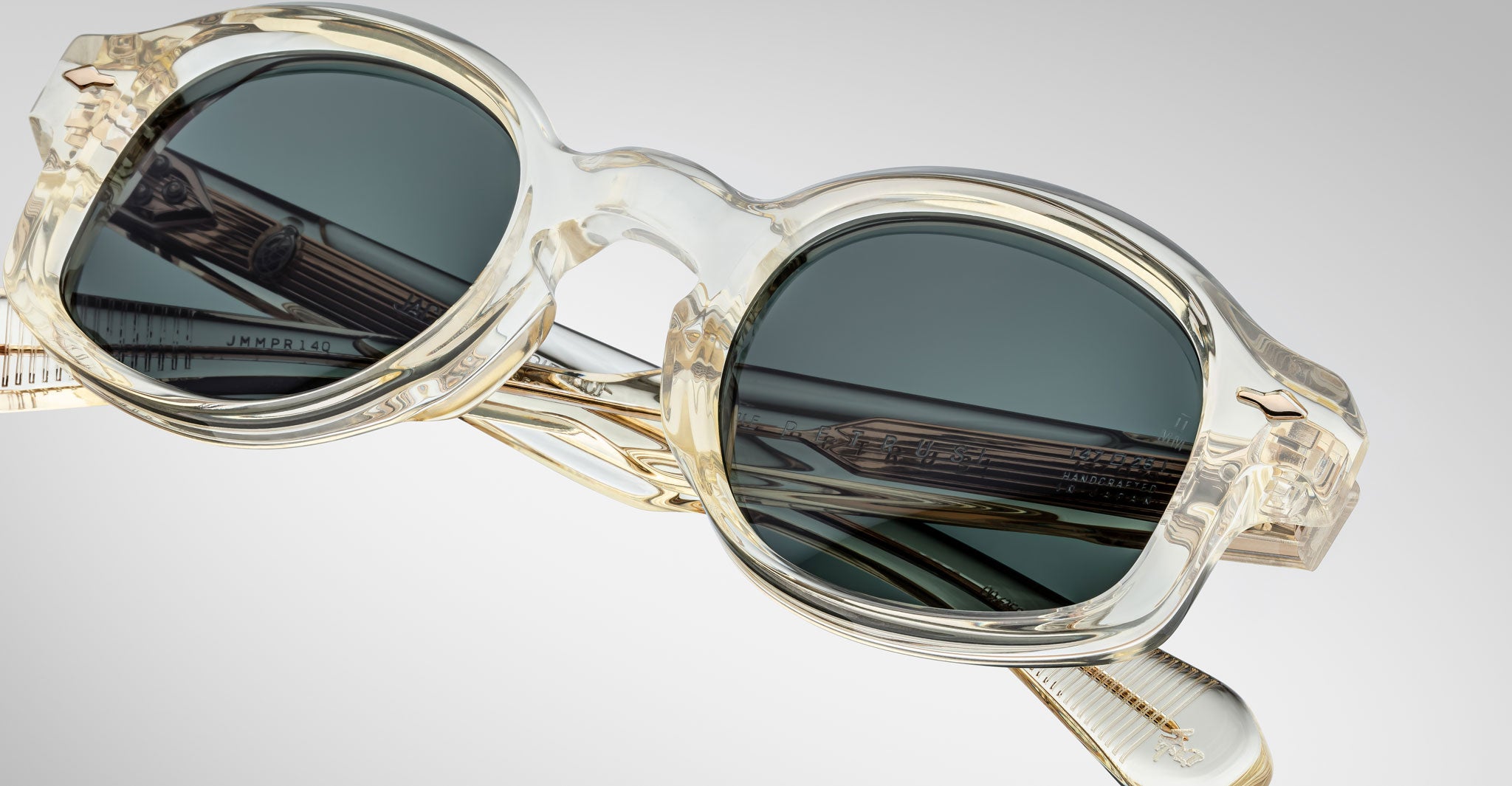 Close-up view of Jacques Marie Mage Petrus round sunglasses featuring transparent acetate frames and dark green lenses, with detailed texture and inner arm engraving, set against a plain light background.