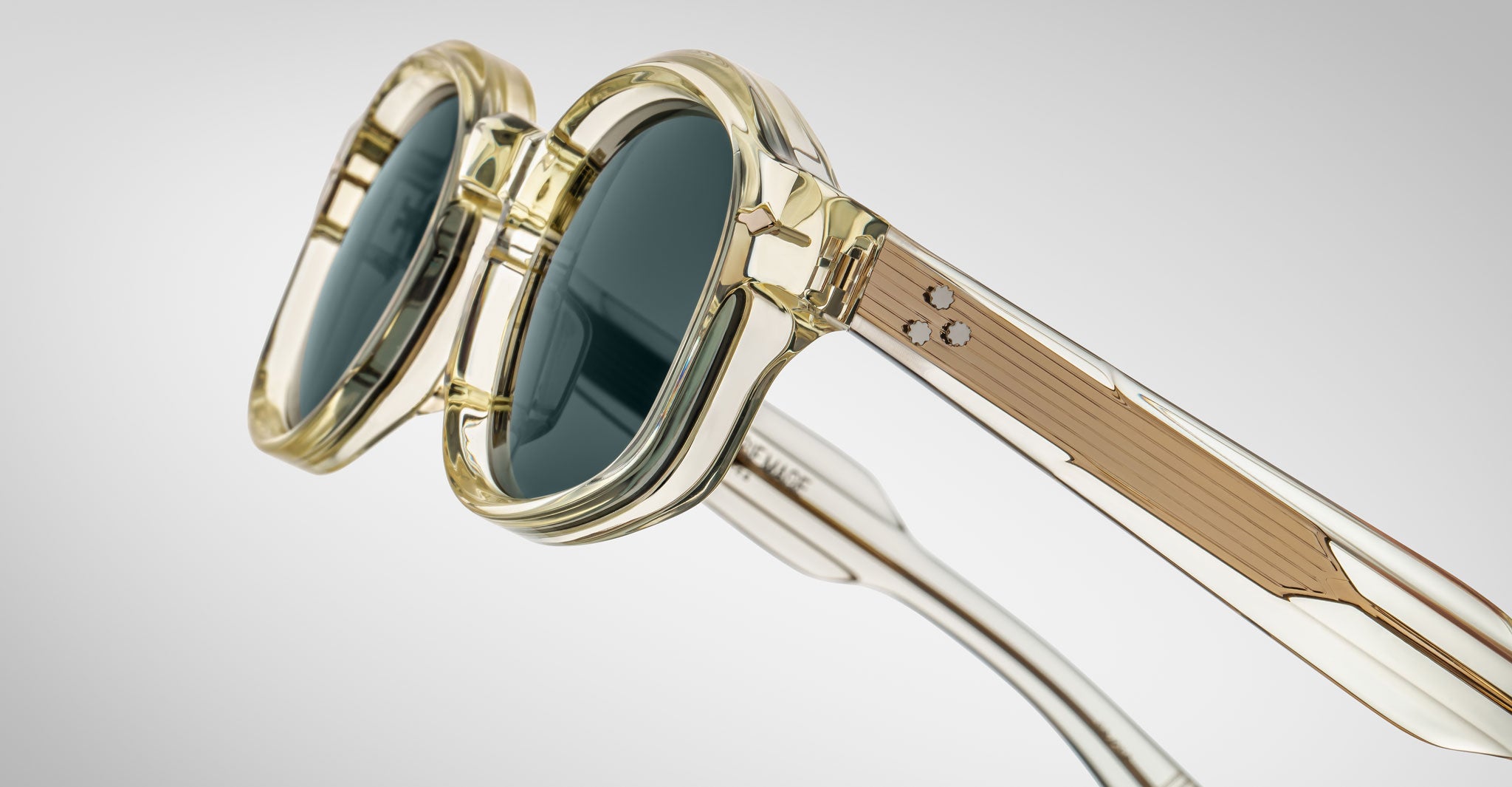 Close-up of Jacques Marie Mage Petrus sunglasses with clear yellowish frames, dark round lenses, and engraved metal temple arms, set against a plain gray gradient background.