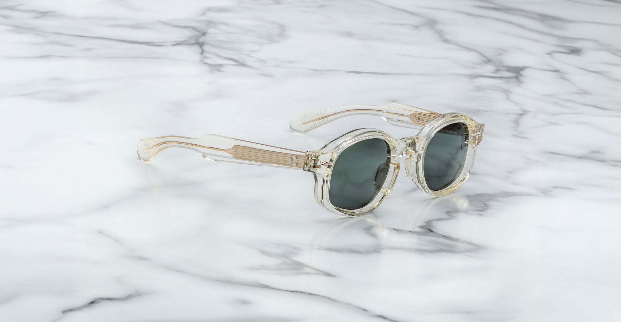 Jacques Marie Mage Petrus sunglasses with clear, round frames and dark lenses are displayed on a white marble surface. The transparent design features subtle beige tones and visible hinges on the temples.