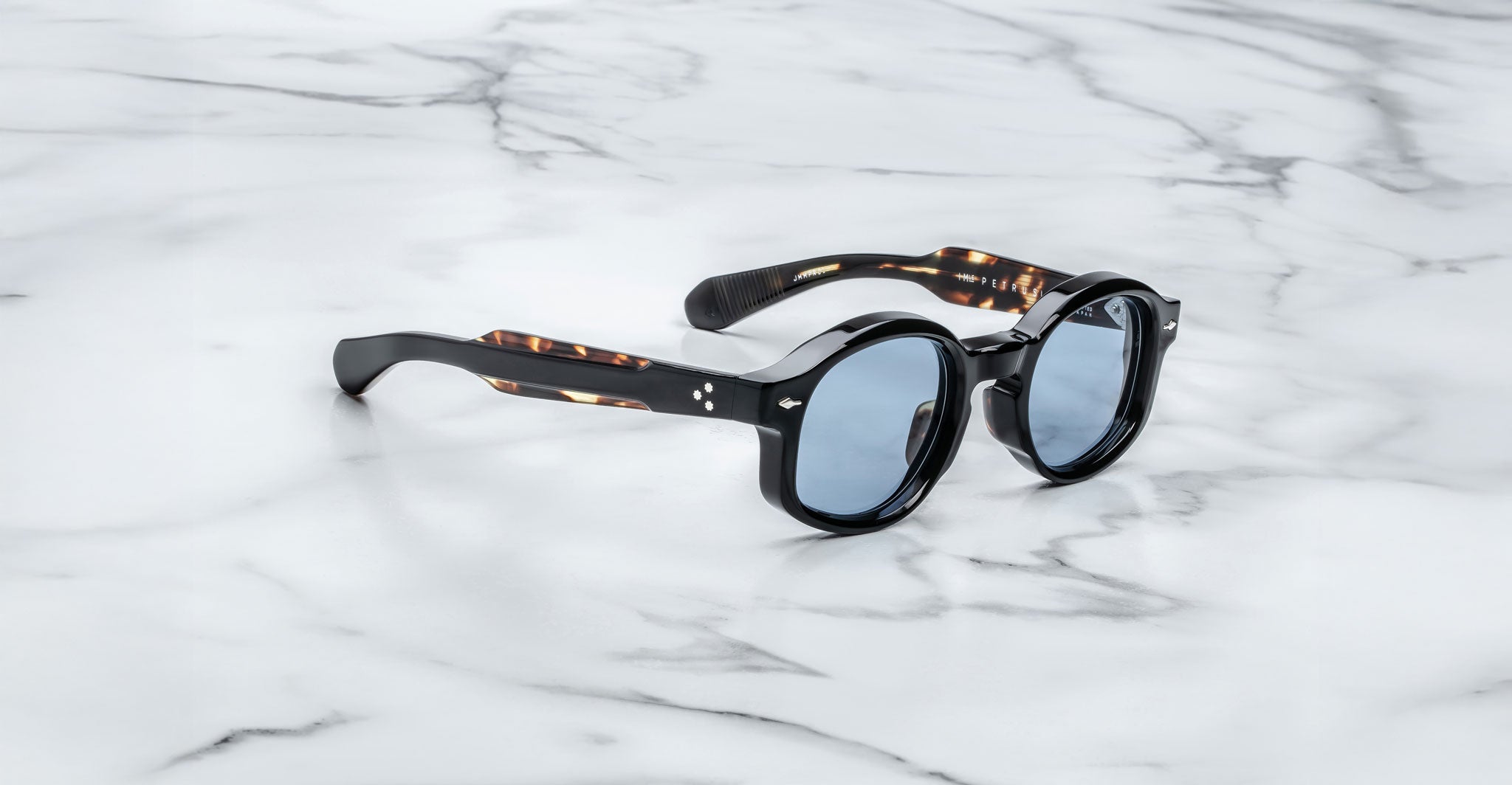 Jacques Marie Mage Petrus round sunglasses with black frames and blue-tinted lenses rest on a white marble surface. The arms feature a subtle tortoiseshell pattern.