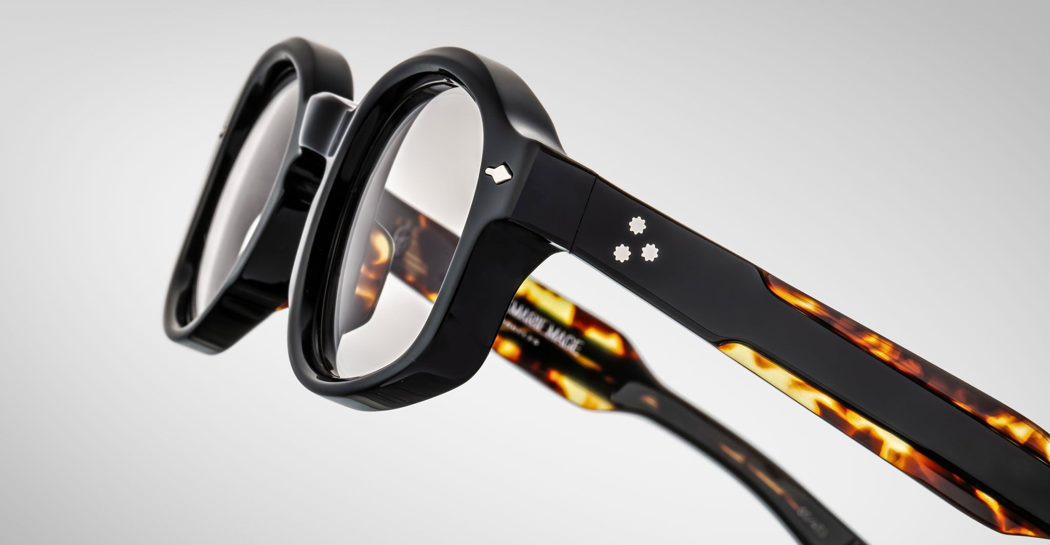Close-up of Jacques Marie Mage Petrus eyeglasses featuring thick black frames, tortoiseshell-patterned arms, and small metallic accents on the temples, set against a plain light gray background.