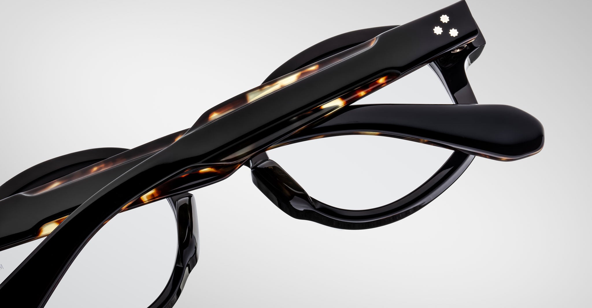 Close-up of two Jacques Marie Mage Petrus eyeglasses with thick, glossy frames—one in black, one in tortoiseshell—stacked atop each other against a plain light background.