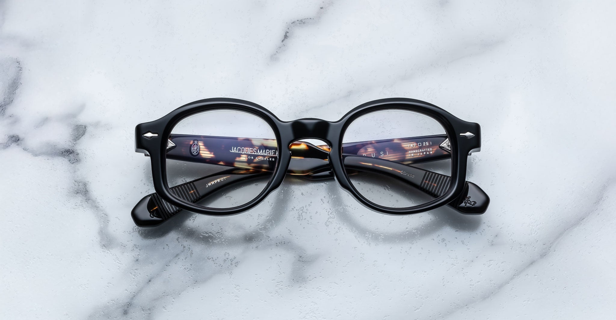 A pair of Jacques Marie Mage Petrus round black eyeglasses lies folded on a white marble surface. The inner arms, partially visible, reveal text and catch a faint reflection of lights.