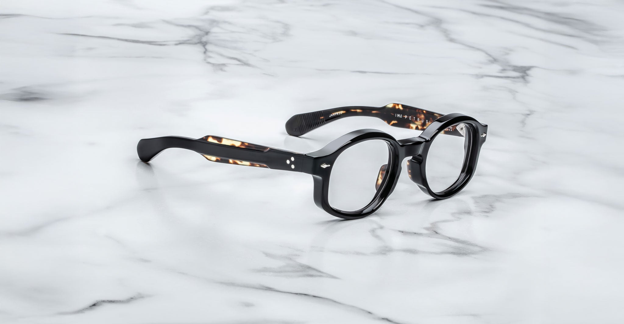 A pair of Jacques Marie Mage Petrus round, black eyeglasses with thick frames and subtly patterned arms rests on a white marble surface with gray veining.