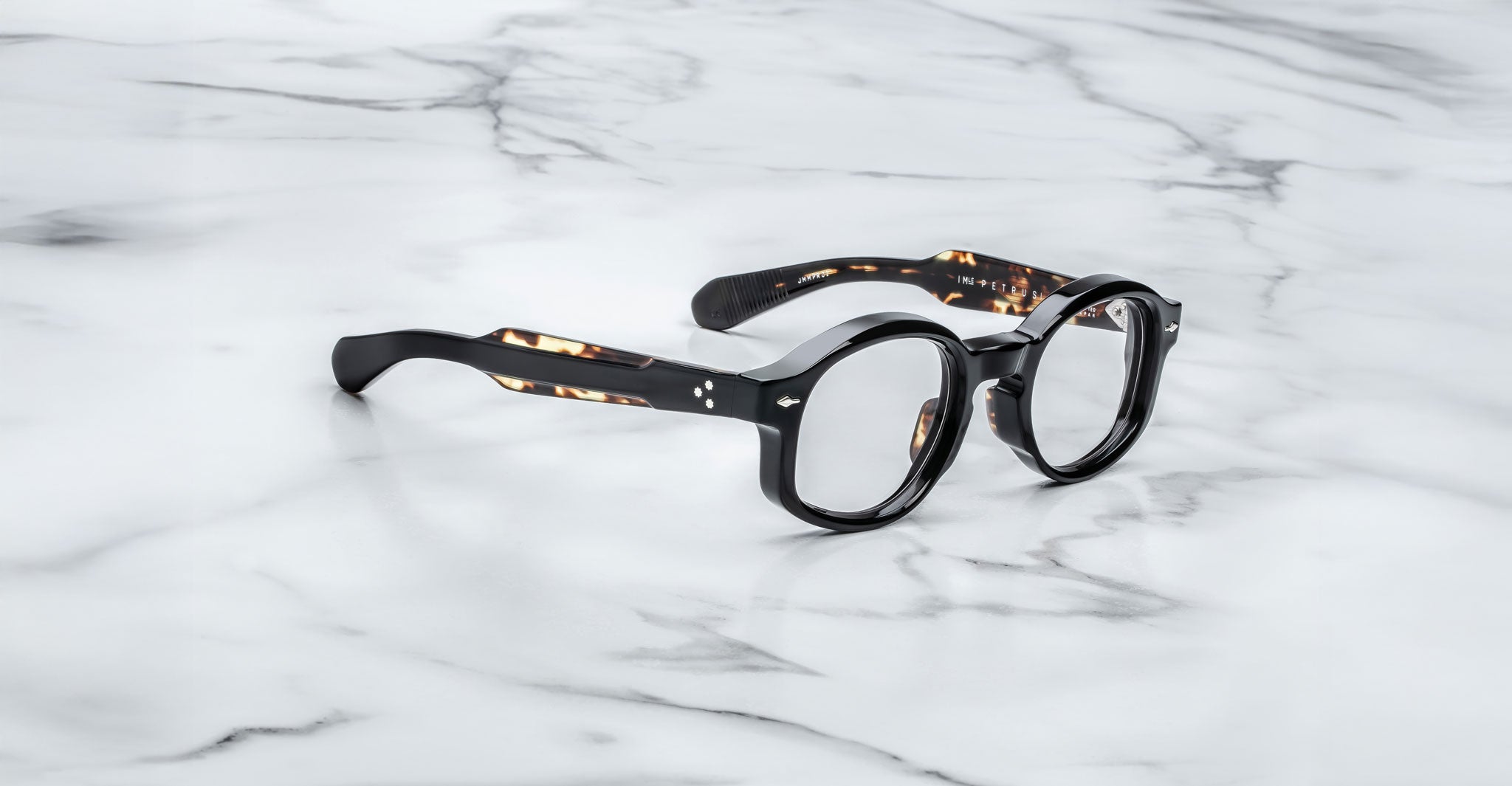 A pair of Jacques Marie Mage Petrus round, black eyeglasses with thick frames and subtly patterned arms rests on a white marble surface with gray veining.