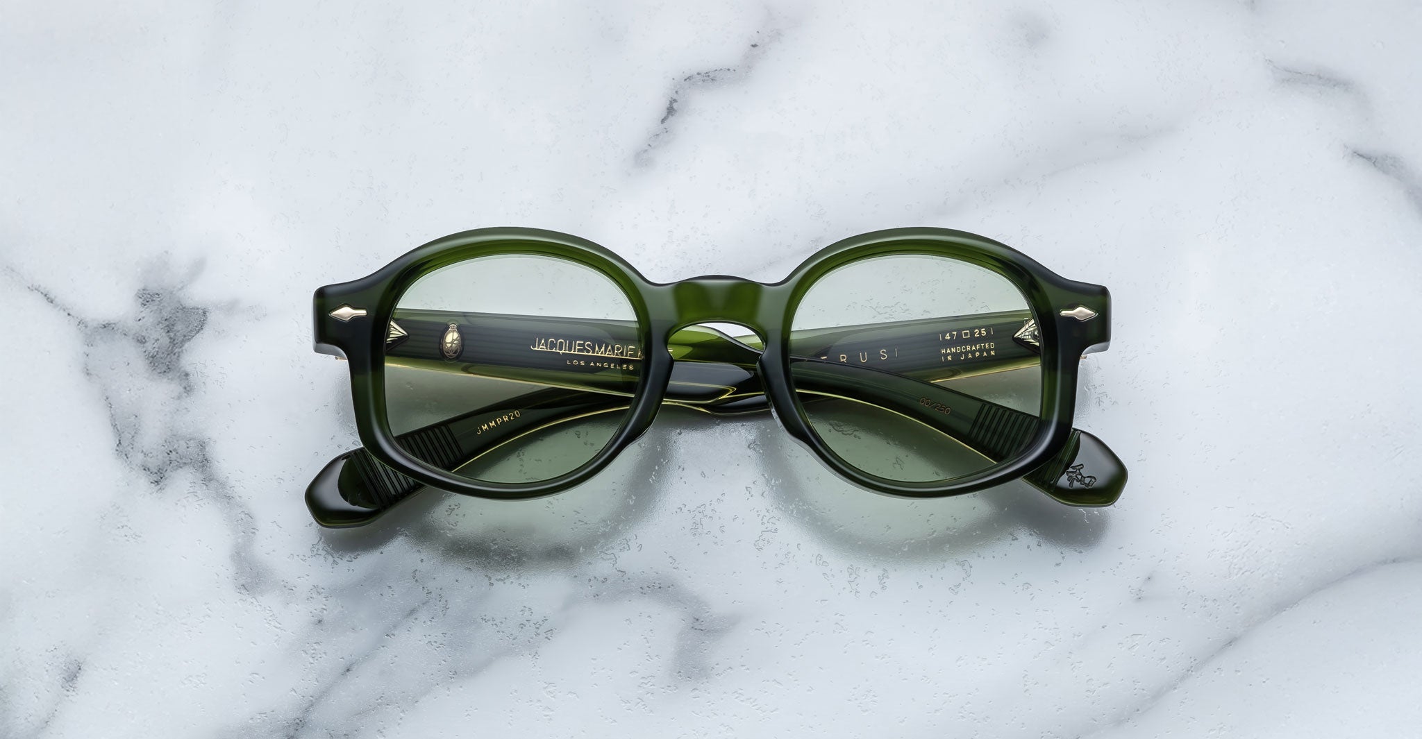 A pair of Jacques Marie Mage Petrus round green eyeglasses with thick frames and slightly green-tinted lenses rests on a white marble surface, their arms neatly folded.