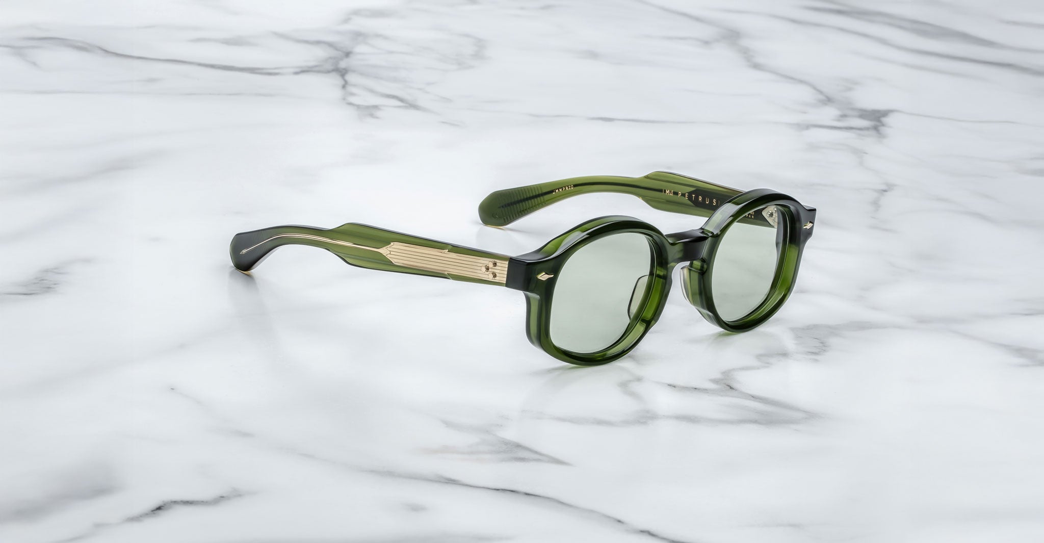 A pair of Jacques Marie Mage Petrus round green sunglasses with gold temple detailing rests on a white marble surface.