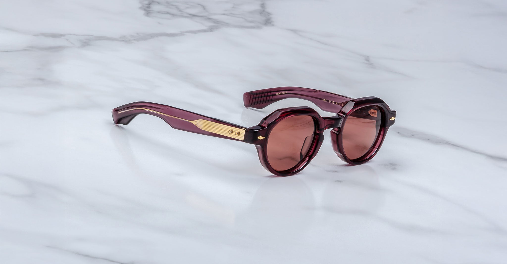 The Jacques Marie Mage Pontian, round dark red sunglasses with gold temple accents, is displayed on a white marble surface.