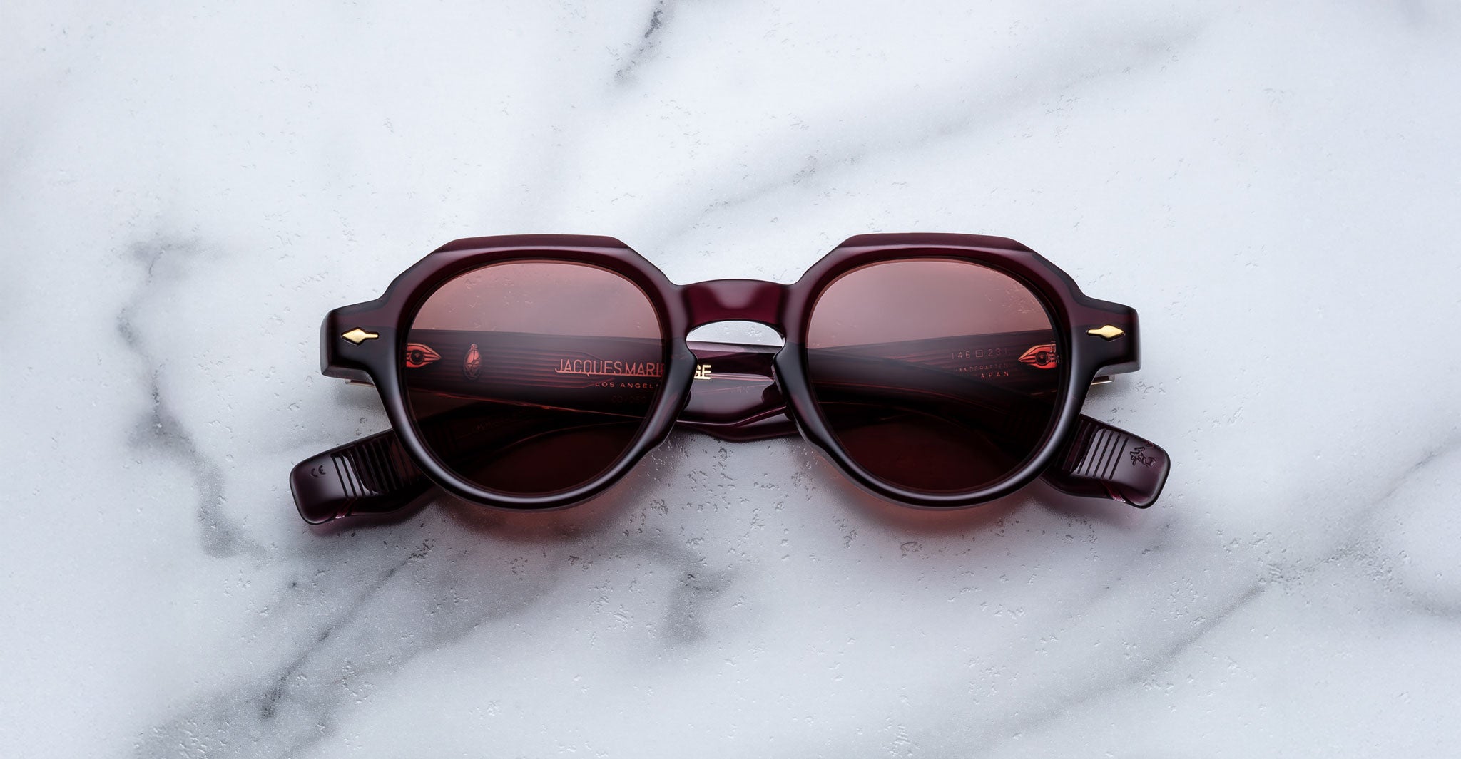 The Jacques Marie Mage Pontian sunglasses, featuring round dark red frames, tinted lenses, and gold arrow accents near the hinges, are displayed on a white marble surface.