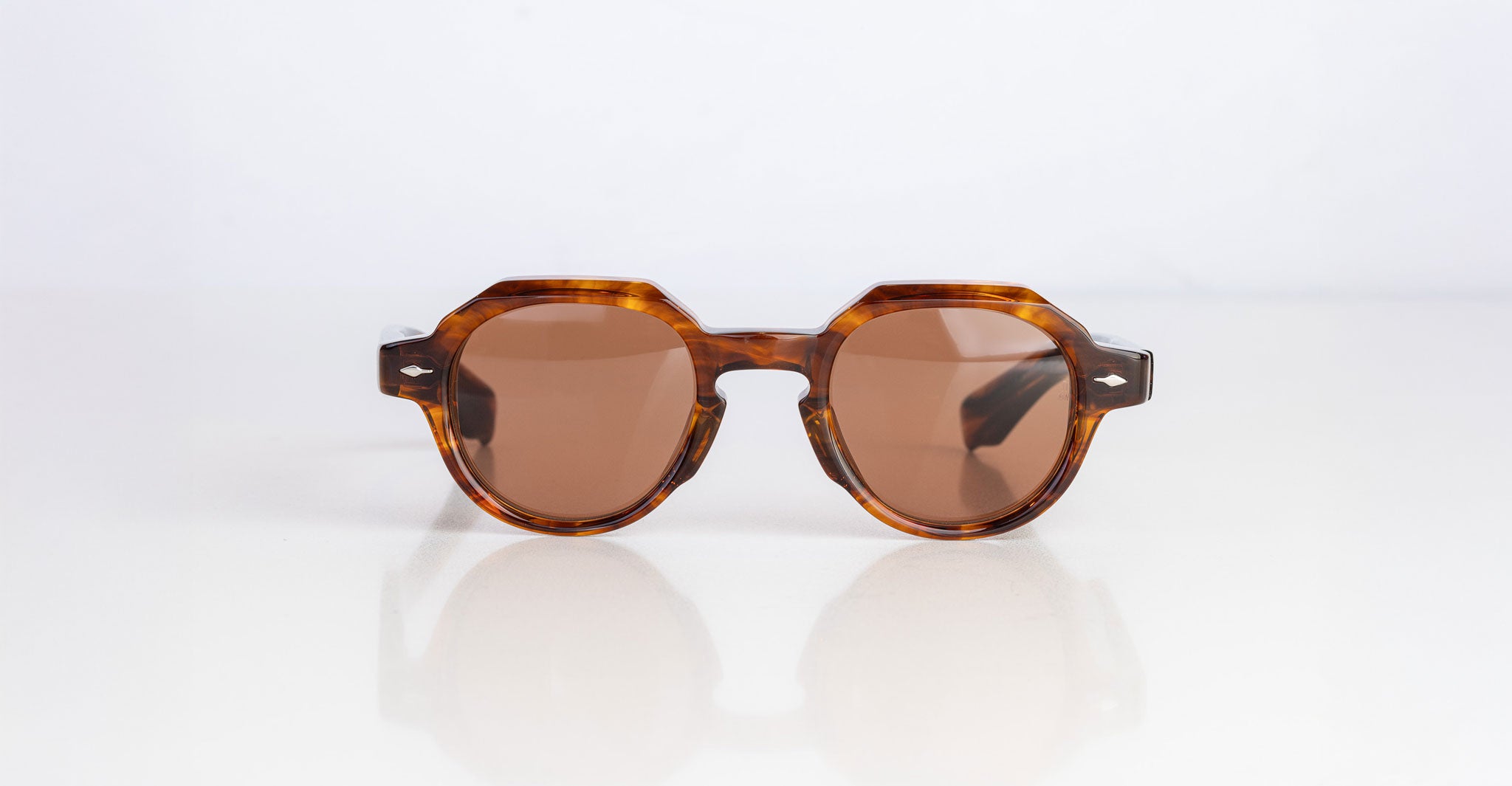 The Jacques Marie Mage Pontian sunglasses feature round, tinted lenses and thick brown tortoiseshell frames, shown on a white surface with a plain white background.
