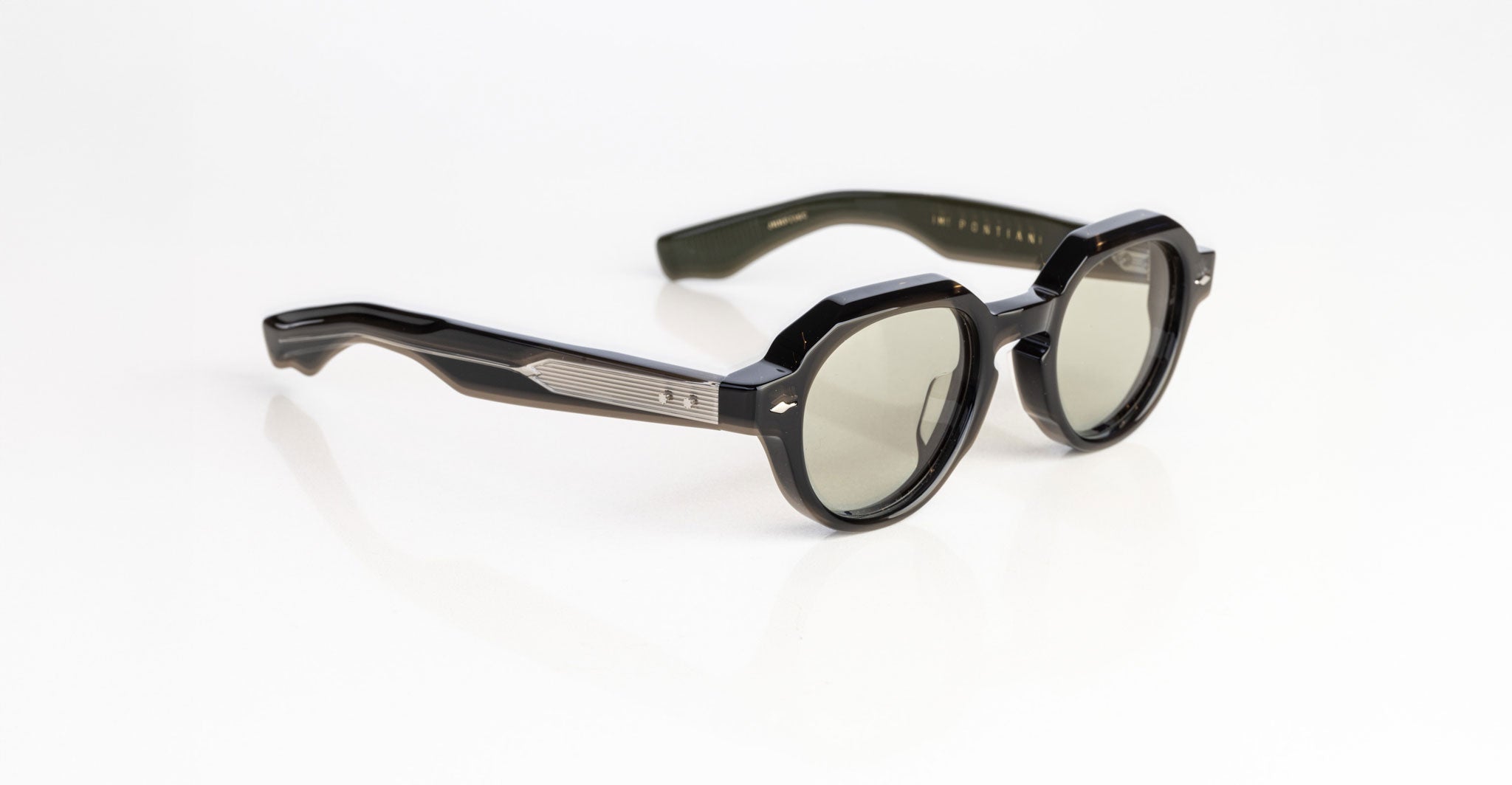 The Jacques Marie Mage Pontian sunglasses feature black, round tinted lenses with thick frames and silver accents by the hinges, displayed on a white surface.
