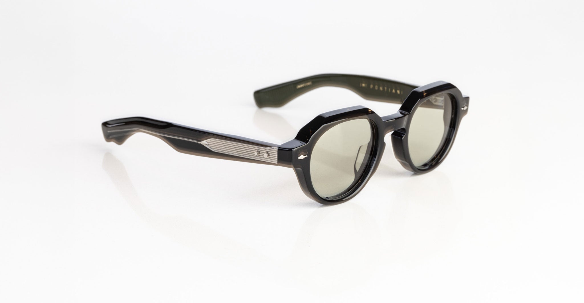 The Jacques Marie Mage Pontian sunglasses feature black, round tinted lenses with thick frames and silver accents by the hinges, displayed on a white surface.