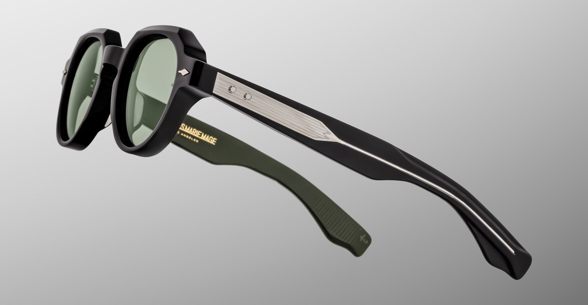 The Jacques Marie Mage Pontian sunglasses feature black frames, green-tinted lenses, metallic detailing on the arms, and a subtle arrow logo near the hinges, all set against a plain gray background.