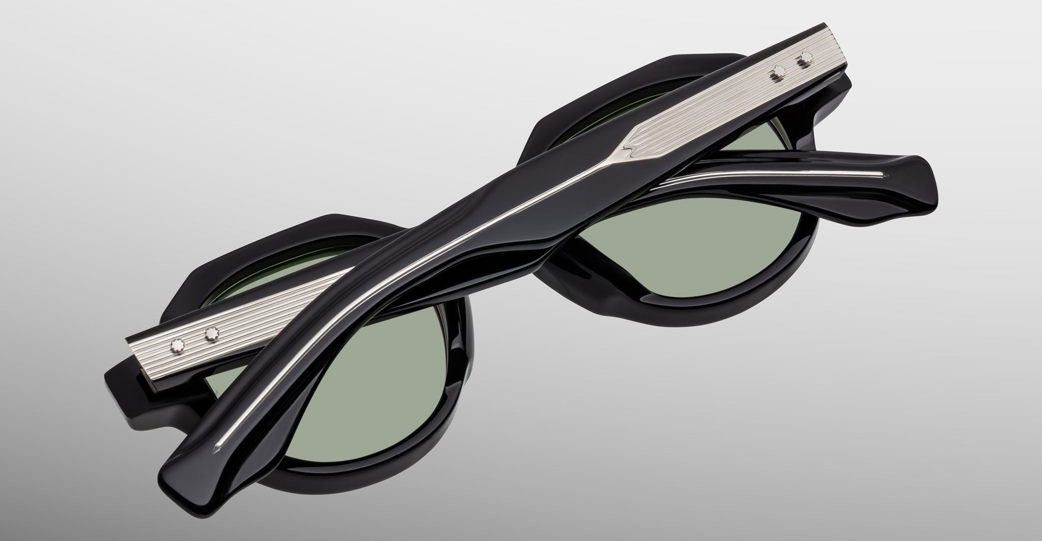 A pair of Jacques Marie Mage Pontian sunglasses in black and silver with green lenses, folded on a light gray surface with the arms crossed over the front of the frames.