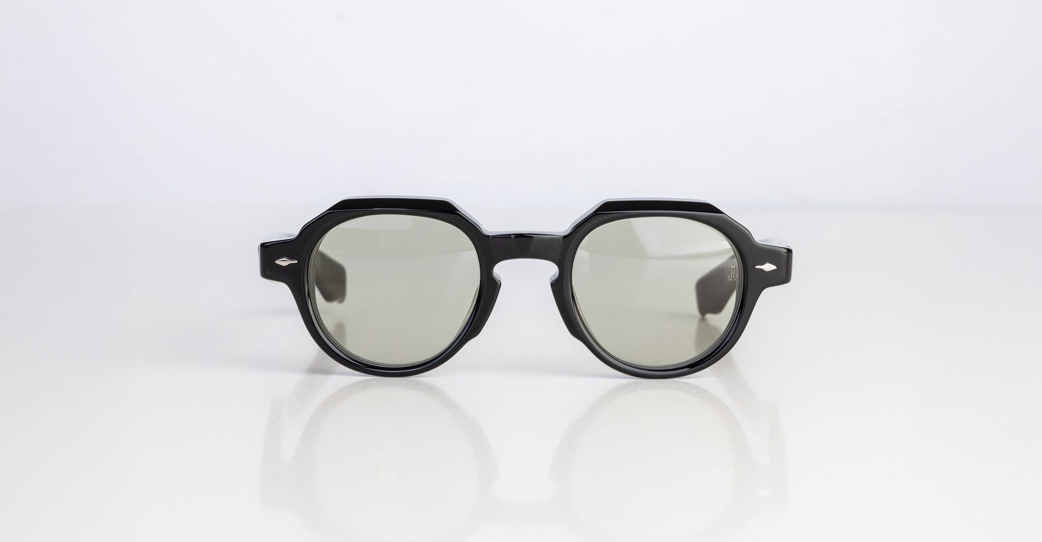 The Jacques Marie Mage Pontian sunglasses, featuring thick geometric black frames and gray tinted lenses, are centered on a reflective white surface against a plain light-colored background.