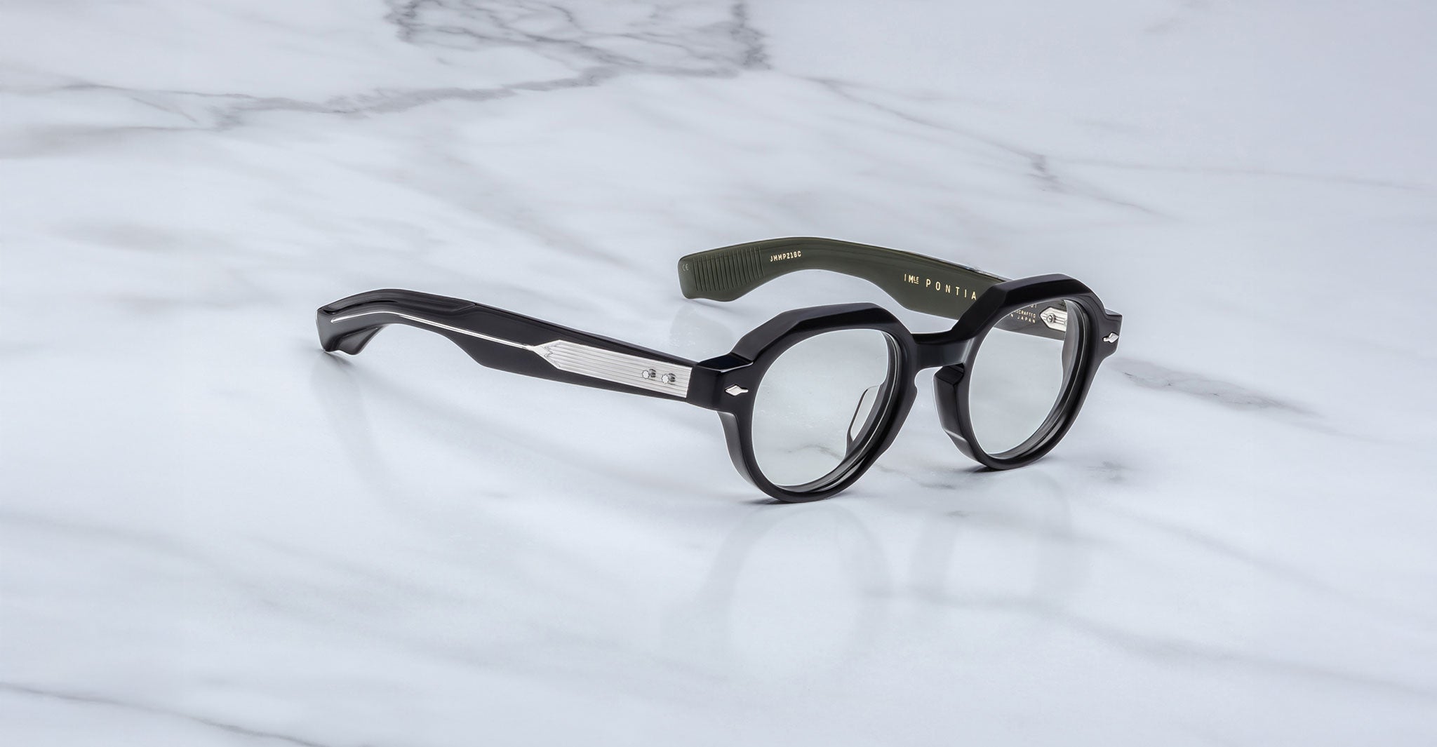 The Jacques Marie Mage Pontian eyeglasses feature round black lenses, thick rims, metallic-accented temples, and an olive green inner arm, displayed on a white marble surface.