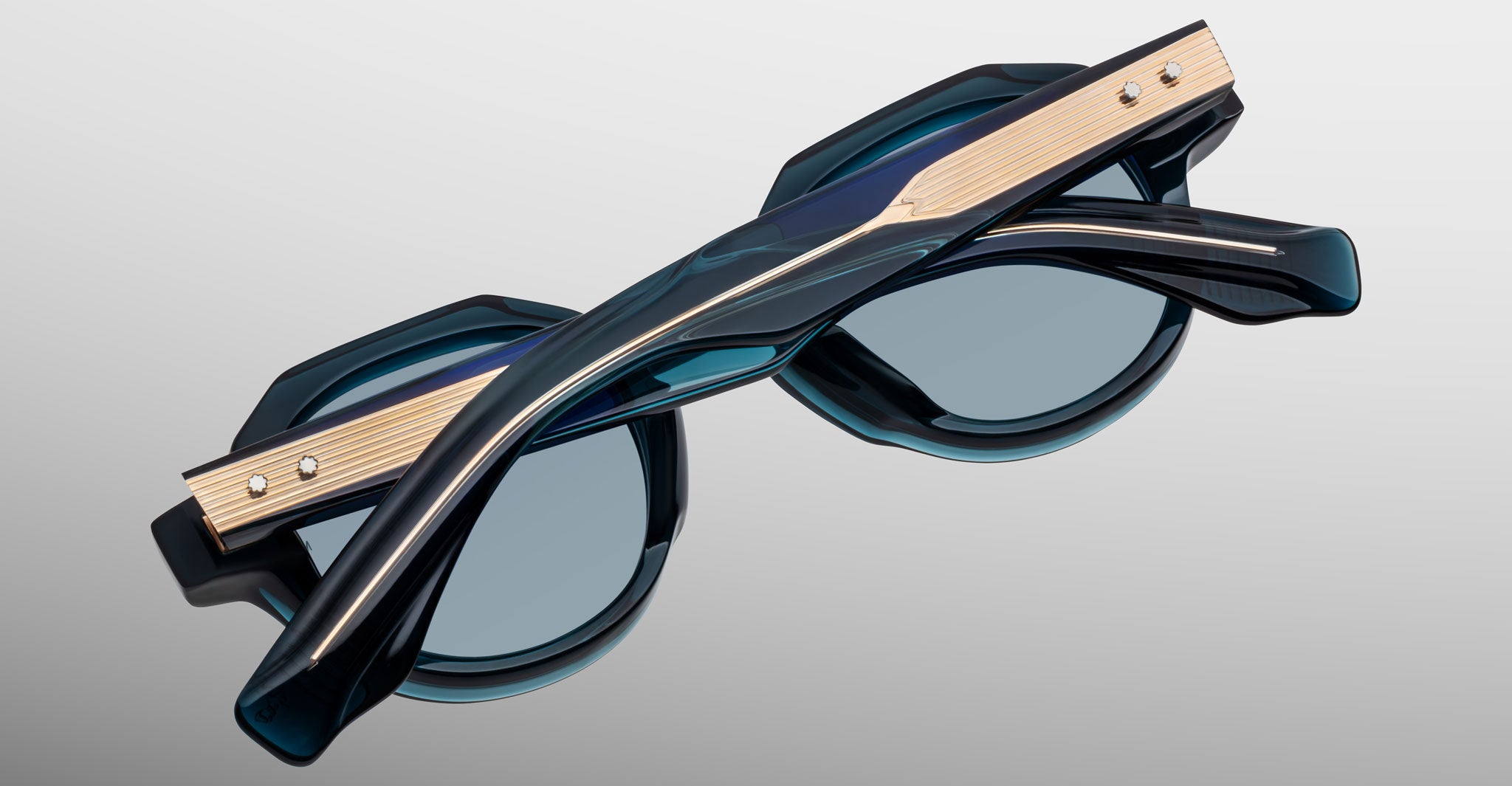 A folded pair of Jacques Marie Mage Pontian sunglasses with dark teal, octagonal lenses and metallic gold temple accents, resting on a light grey background.