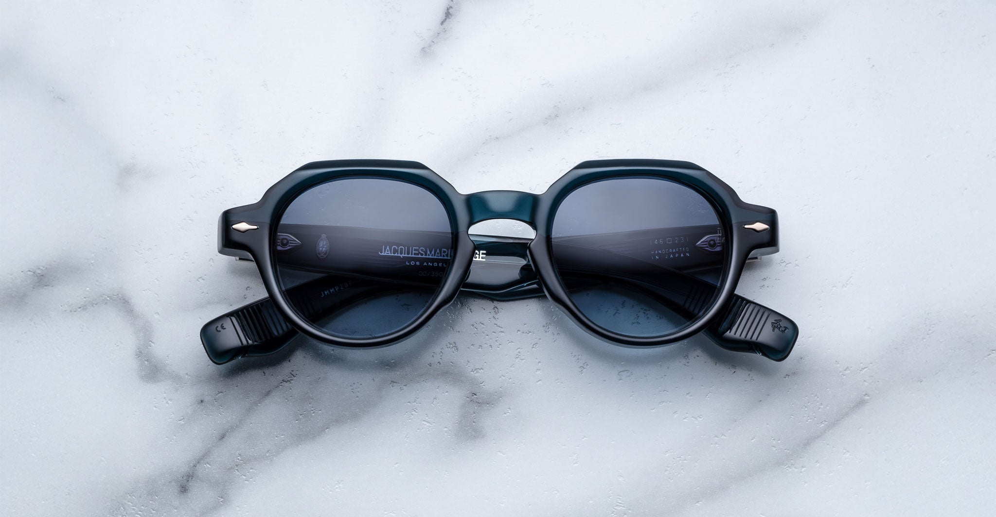 The Jacques Marie Mage Pontian sunglasses, featuring round dark lenses and thick black frames, are displayed on a white marble surface with folded arms revealing the interior of the frames.