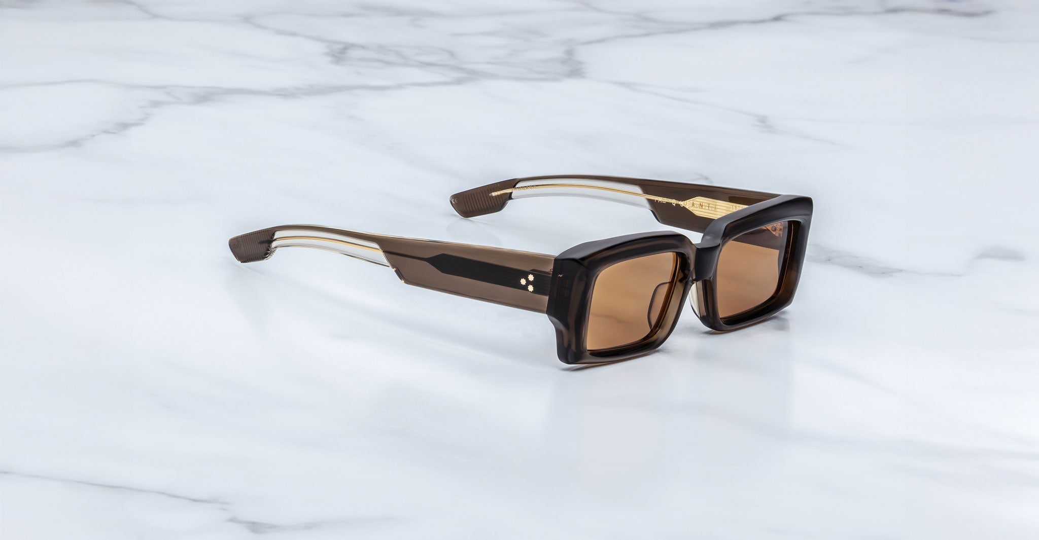 Jacques Marie Mage Quant in London, rectangular translucent brown acetate sunglasses shown from a side angle, featuring bold sculpted rims, wide temples with signature metal inlay, and warm brown tinted lenses on a light marble surface.