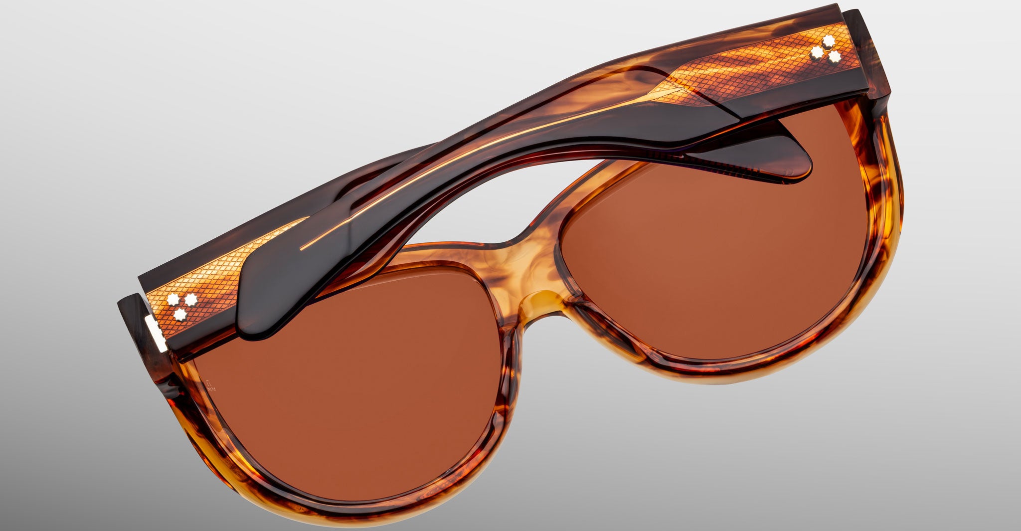 The Jacques Marie Mage Ravello sunglasses, featuring a brown tortoiseshell frame and orange-tinted lenses, are shown folded with the arms crossing over the front on a light gray background.