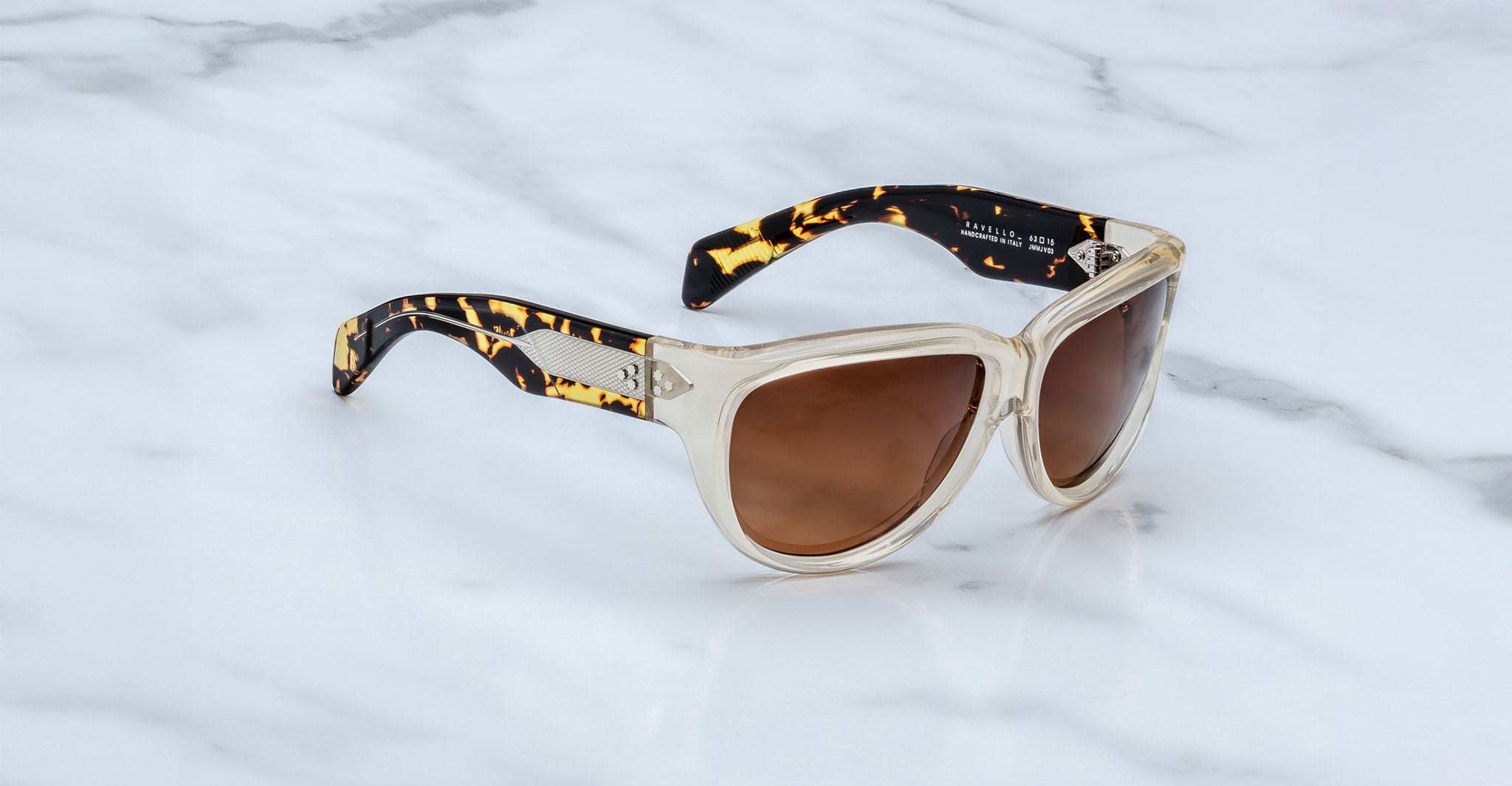 The Jacques Marie Mage Ravello sunglasses feature transparent frames, brown-tinted lenses, and tortoiseshell-patterned arms, displayed on a white marble surface.