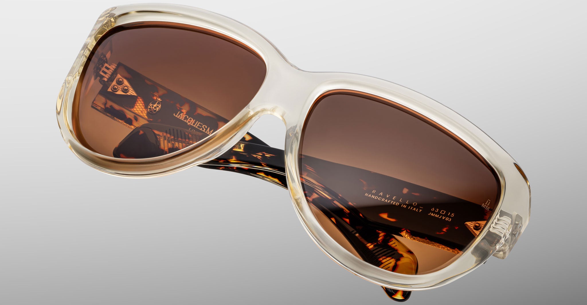 A close-up of the Jacques Marie Mage Ravello sunglasses featuring clear oversized frames, brown tinted lenses, and tortoiseshell-patterned arms, displayed against a plain light gray background.