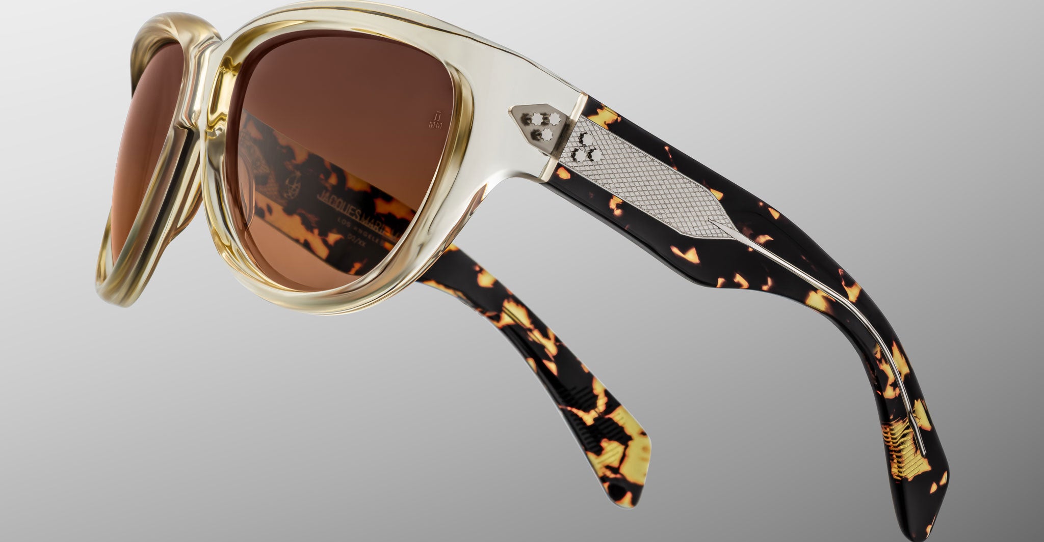 Close-up of Jacques Marie Mage Ravello sunglasses with translucent beige frames, brown tinted lenses, and black and yellow tortoiseshell-patterned arms with metal detailing by the hinges, set against a neutral gradient background.