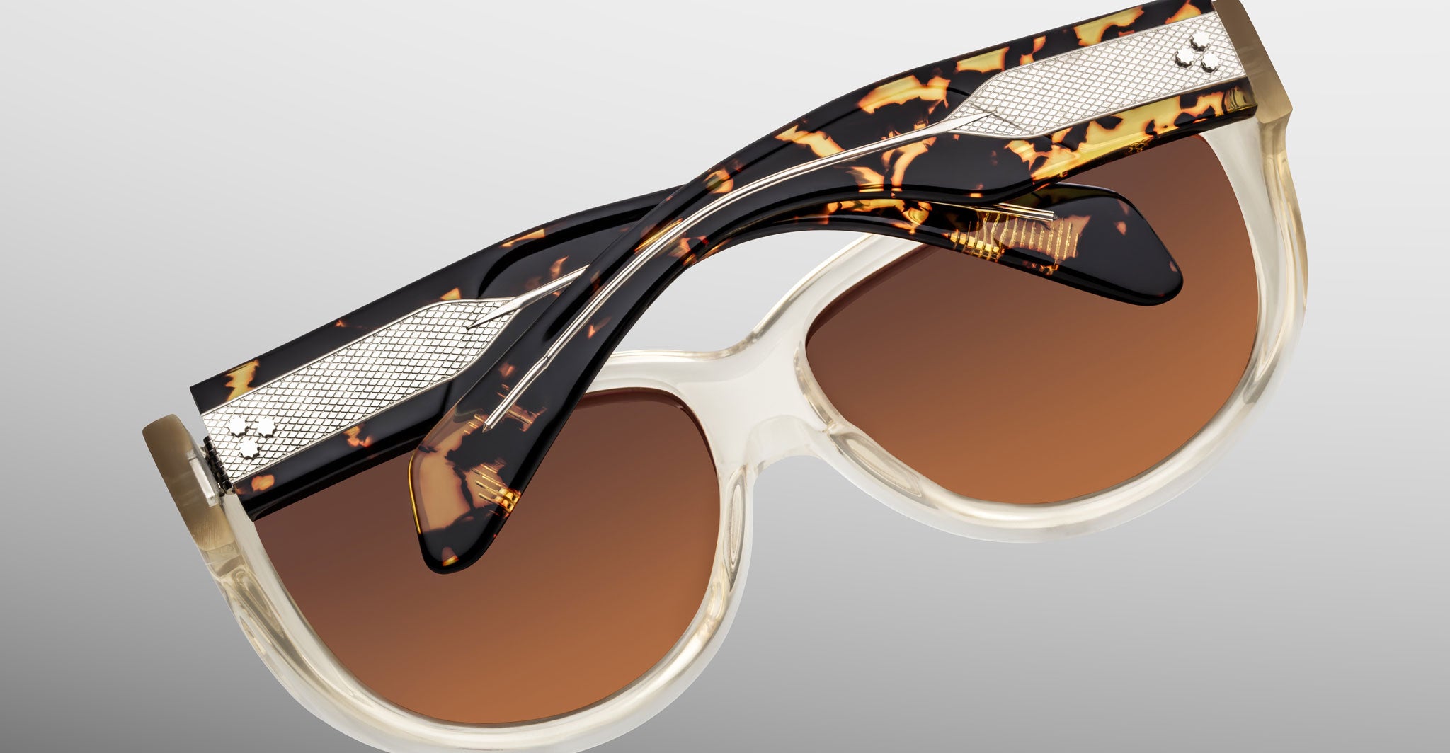 Close-up of Jacques Marie Mage Ravello sunglasses with brown-tinted lenses, clear rims, tortoiseshell-patterned arms, textured metal accents, and gold details, set against a neutral gradient background.