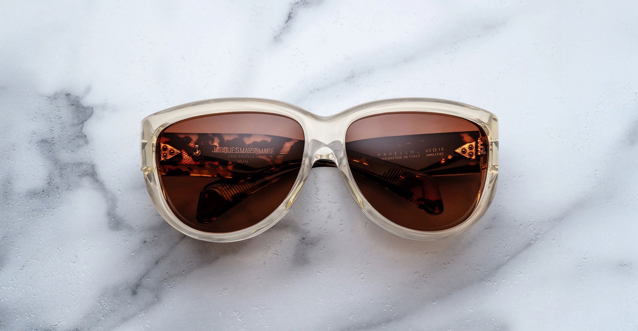 A pair of Jacques Marie Mage Ravello sunglasses with oversized translucent beige frames and brown tinted lenses rests on a white marble surface, with text visible on the inside of the arms.