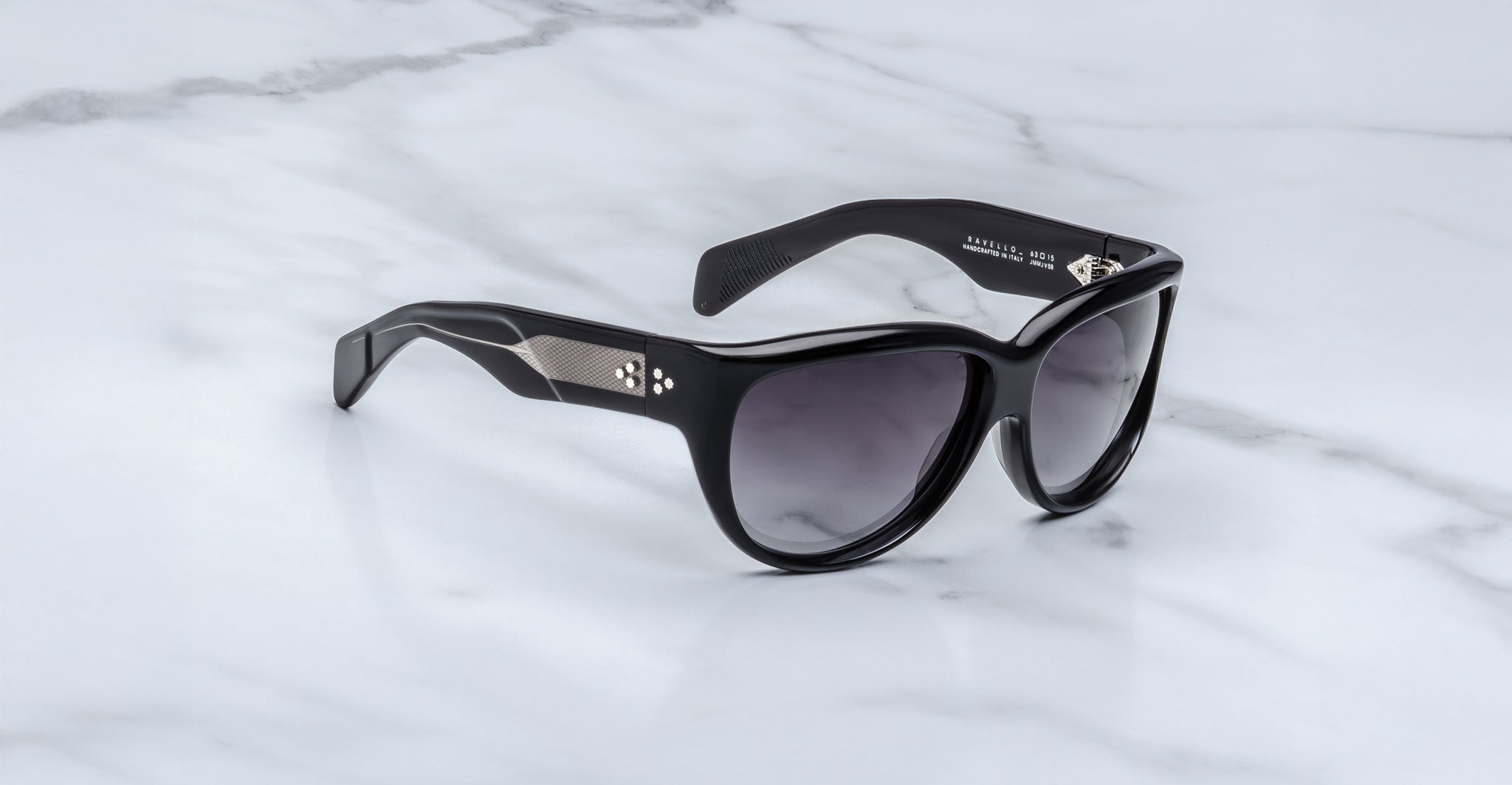 The Jacques Marie Mage Ravello sunglasses, featuring dark lenses, thick frames, and temples adorned with silver metal diamond-shaped detailing, are displayed on a white marble surface.