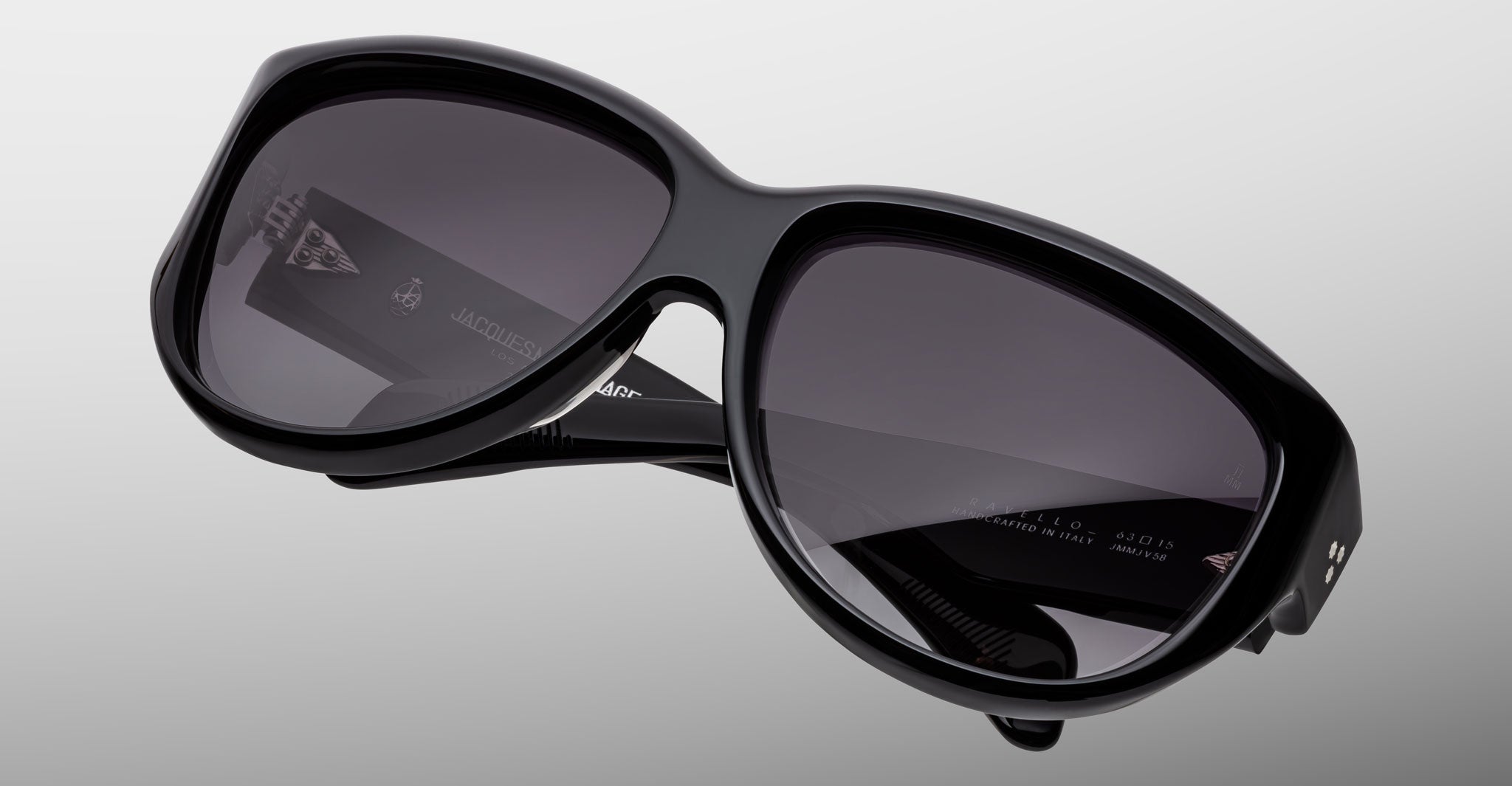 A close-up of black Jacques Marie Mage Ravello sunglasses with dark lenses, displayed against a plain gray background. The Jacques Marie Mage logo is visible on the left lens and the inside of the left temple arm.