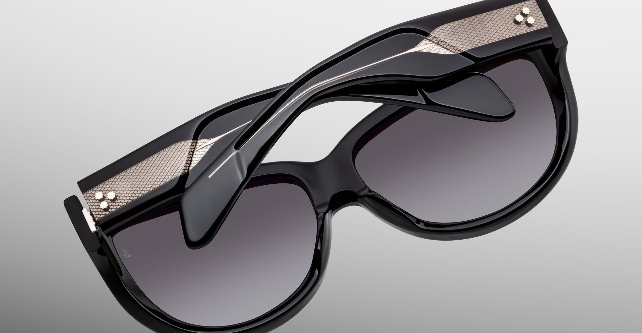 The Jacques Marie Mage Ravello sunglasses feature black frames, dark gradient lenses, metallic gold accents, and layered sculptural temples, shown against a plain gray background.