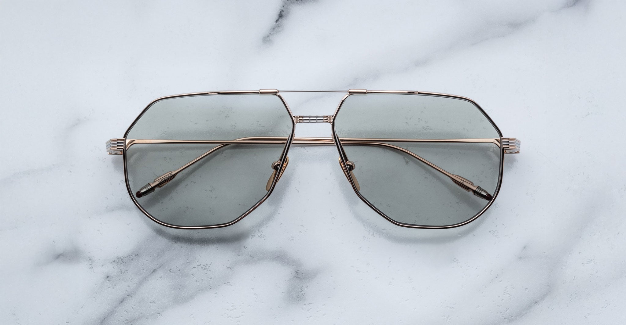 The Jacques Marie Mage Reynold, oversized aviator sunglasses with gold titanium frames and green-tinted lenses inspired by vintage Americana style, are displayed on a white marble surface.