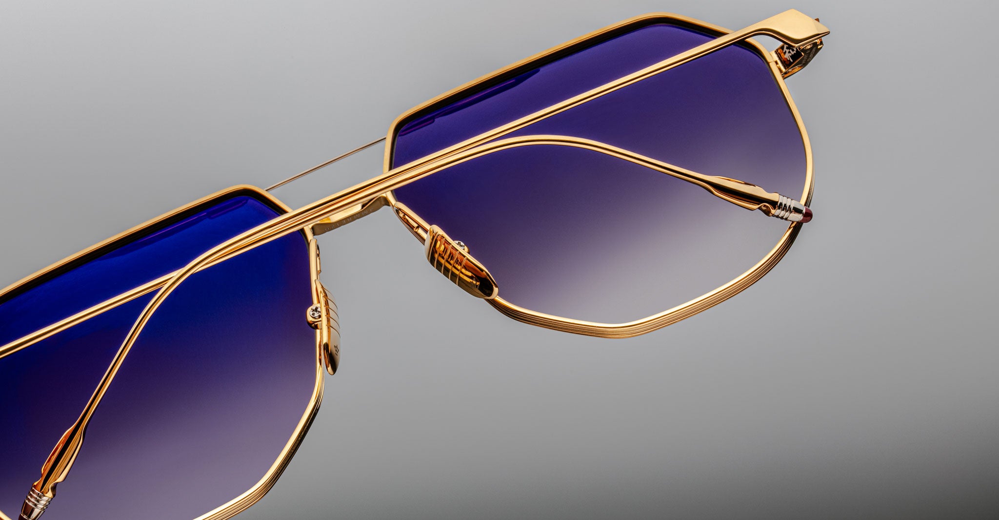 Close-up of Jacques Marie Mage Reynold: stylish oversized aviator sunglasses with a gold titanium frame and blue gradient lenses, featuring a modern geometric design inspired by 70s eyewear, set against a neutral grey background.