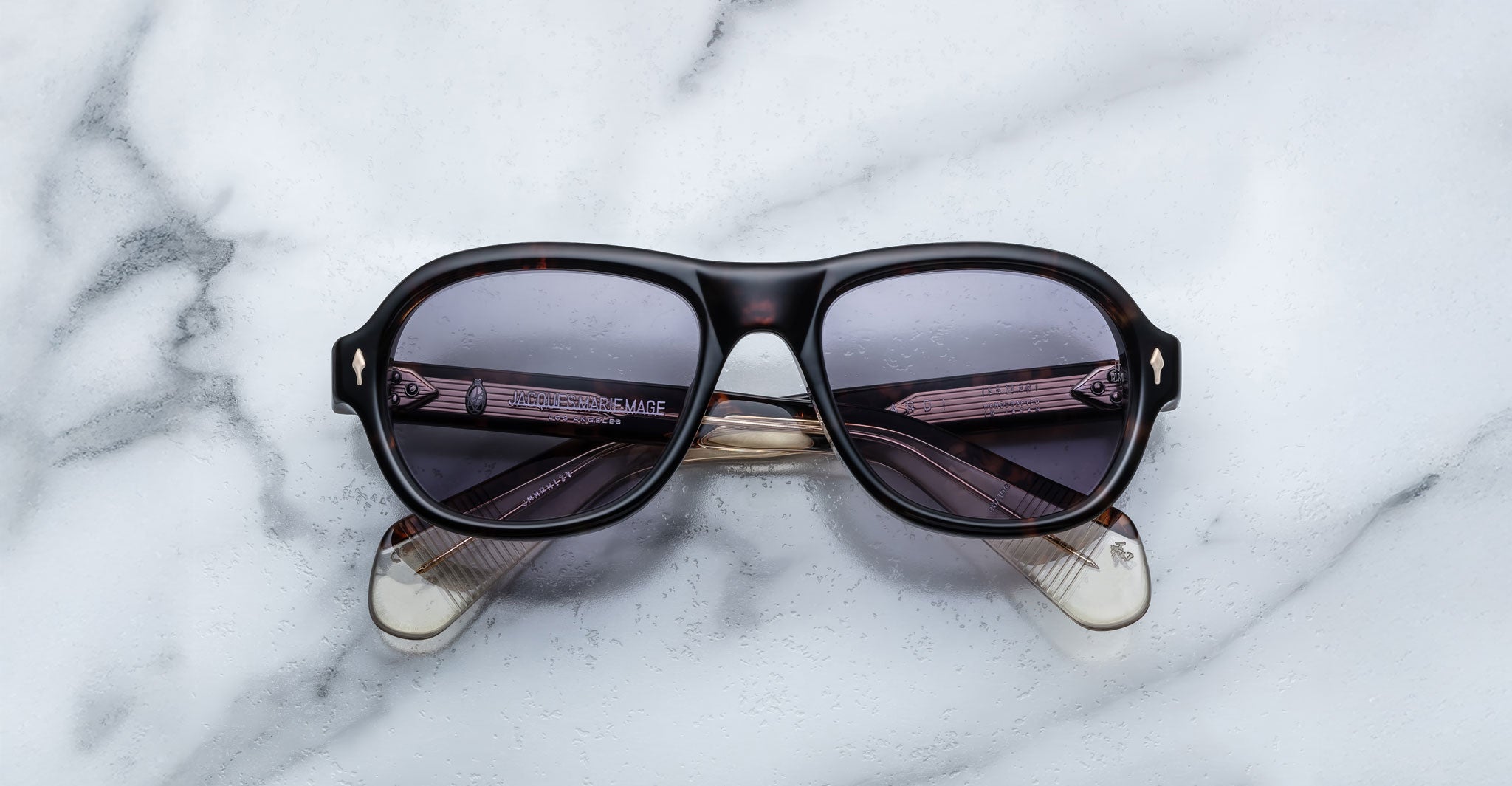 A pair of Jacques Marie Mage Richard Havana, thick-framed sunglasses with blue lenses rests on a white marble surface with grey veins, with faint brand details visible on the inside of the left lens.