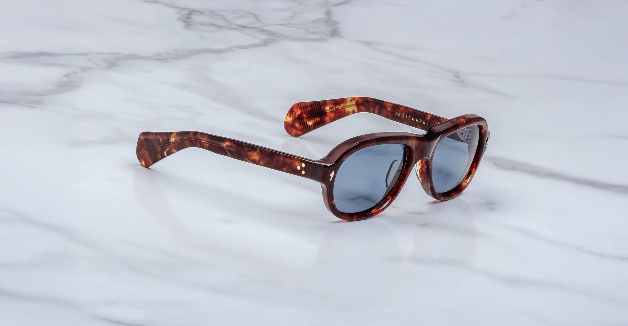 Jacques Marie Mage Richard sunglasses with tortoiseshell thick frames and blue-tinted lenses rest on a white marble surface.
