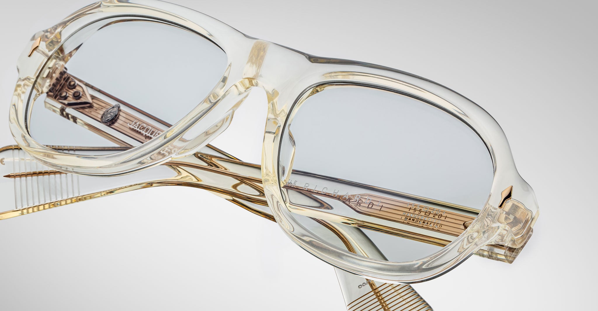 A close-up of Jacques Marie Mage Richard translucent, clear-rimmed eyeglasses featuring gold accents and engraved arm detailing, set against a plain light-gray background.