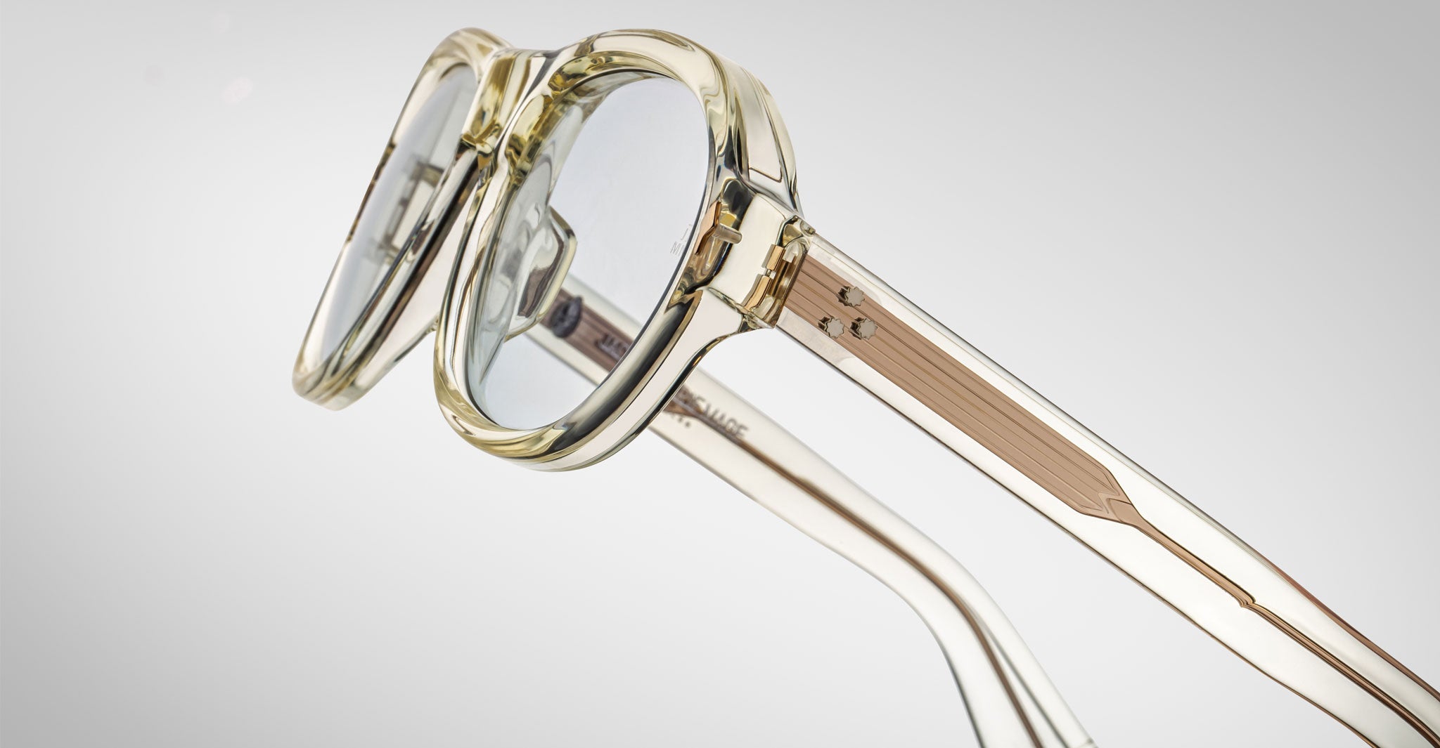 A close-up of Jacques Marie Mage Richard clear round eyeglasses with transparent frames and light gold-tone metal arms, set against a plain light gray background.