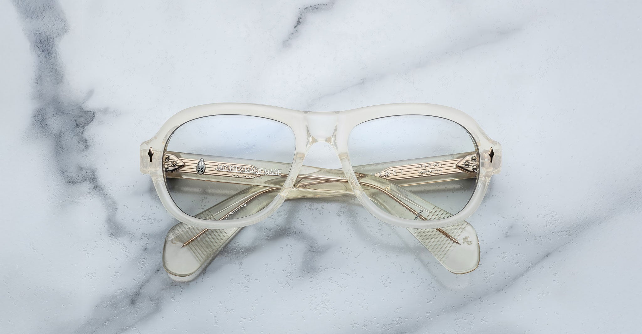 The Jacques Marie Mage Richard eyeglasses, featuring clear frames with a slightly frosted finish, are displayed closed on a white marble surface with the arms crossed behind the lenses.