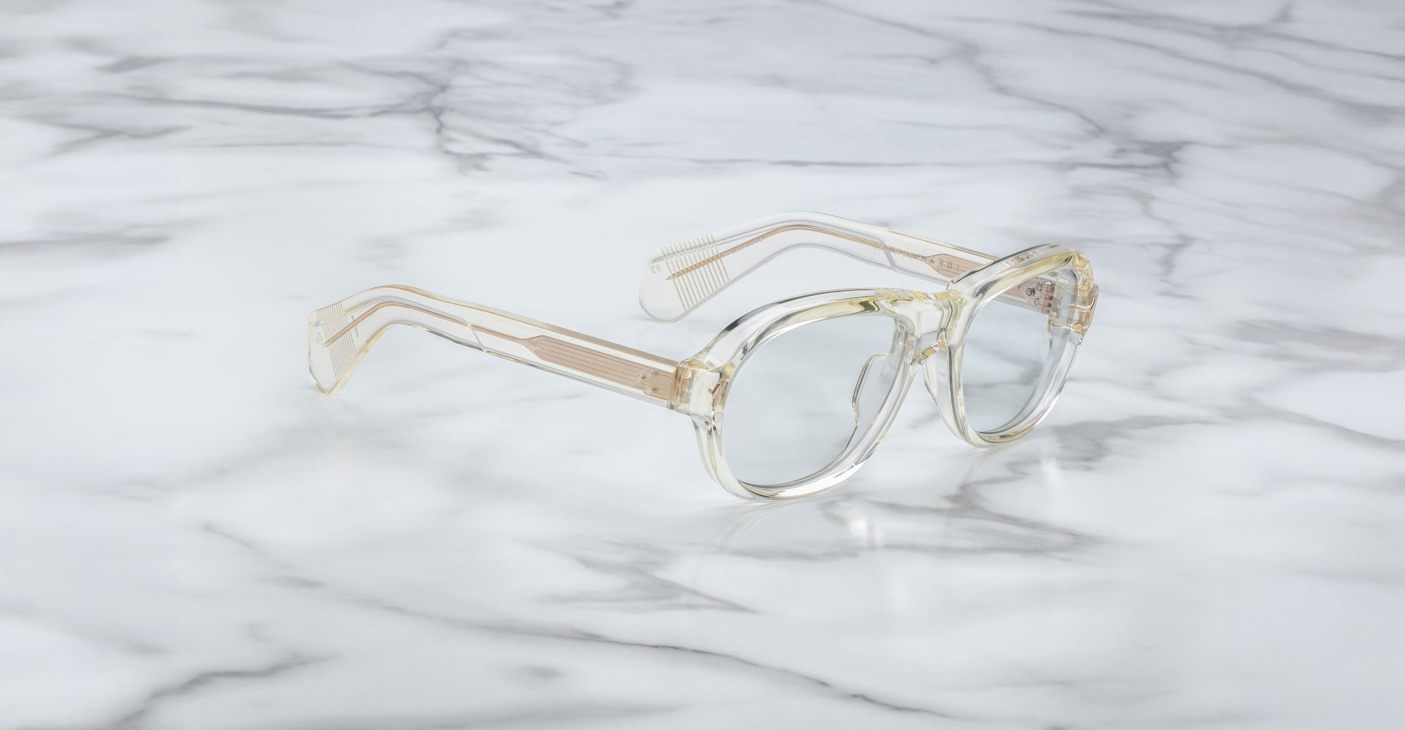 The Jacques Marie Mage Richard eyeglasses, featuring clear thick frames and subtly yellow-tinted temples, rest on a white marble surface with grey veins.